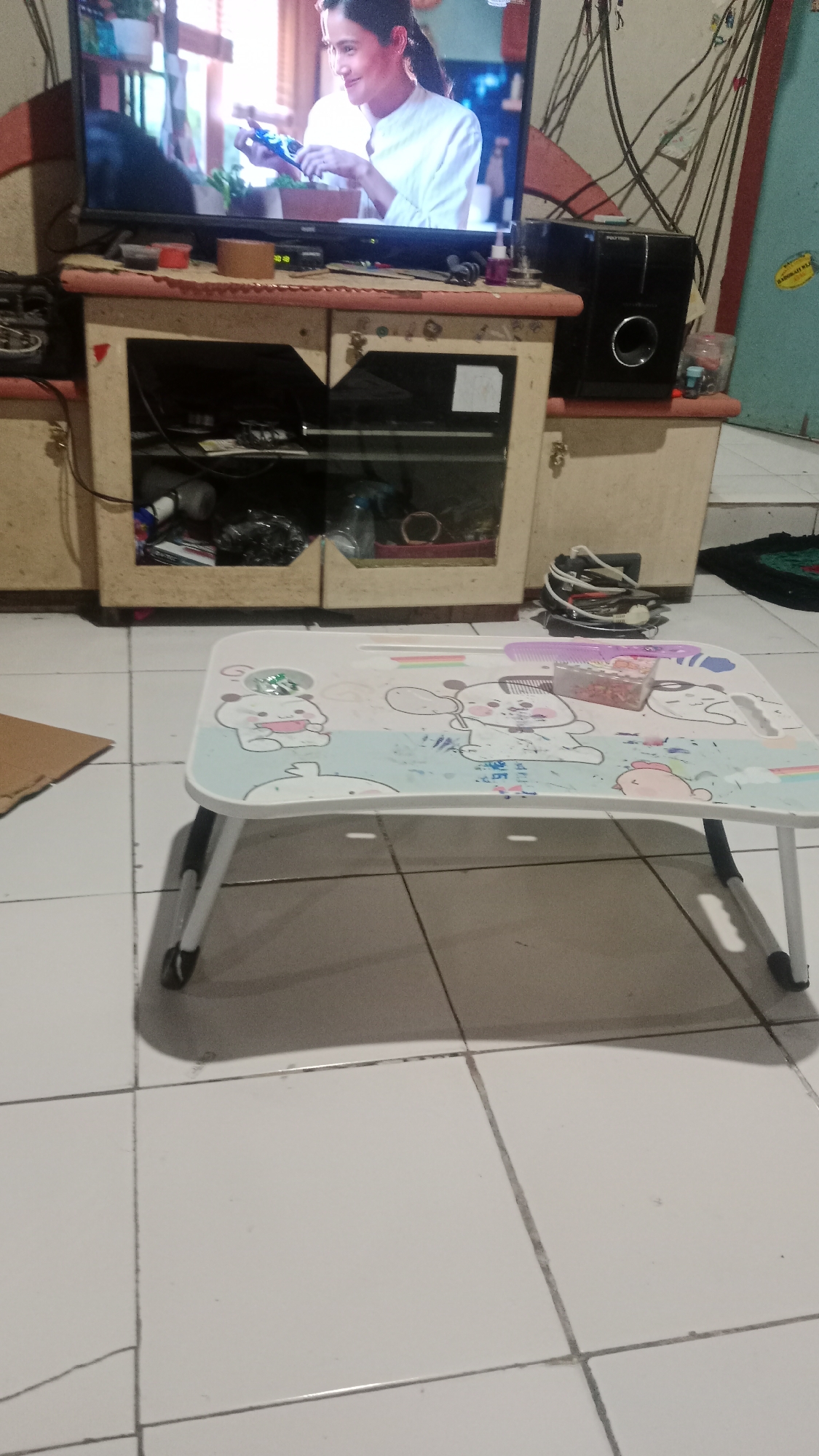 The image shows a portable table with foldable legs placed on a tiled floor. The table has a white surface decorated with cartoonish animal illustrations and a small pink comb and a small transparent box containing small colorful items. In the background, a television set is visible, displaying a woman in a white shirt holding something. To the right of the television, there is a black speaker. Below the television is a wooden cabinet with glass doors, revealing various items stored inside, including bottles and other miscellaneous objects. The overall environment appears to be an indoor setting, possibly a living room. The location is identified as Blitar, Indonesia.