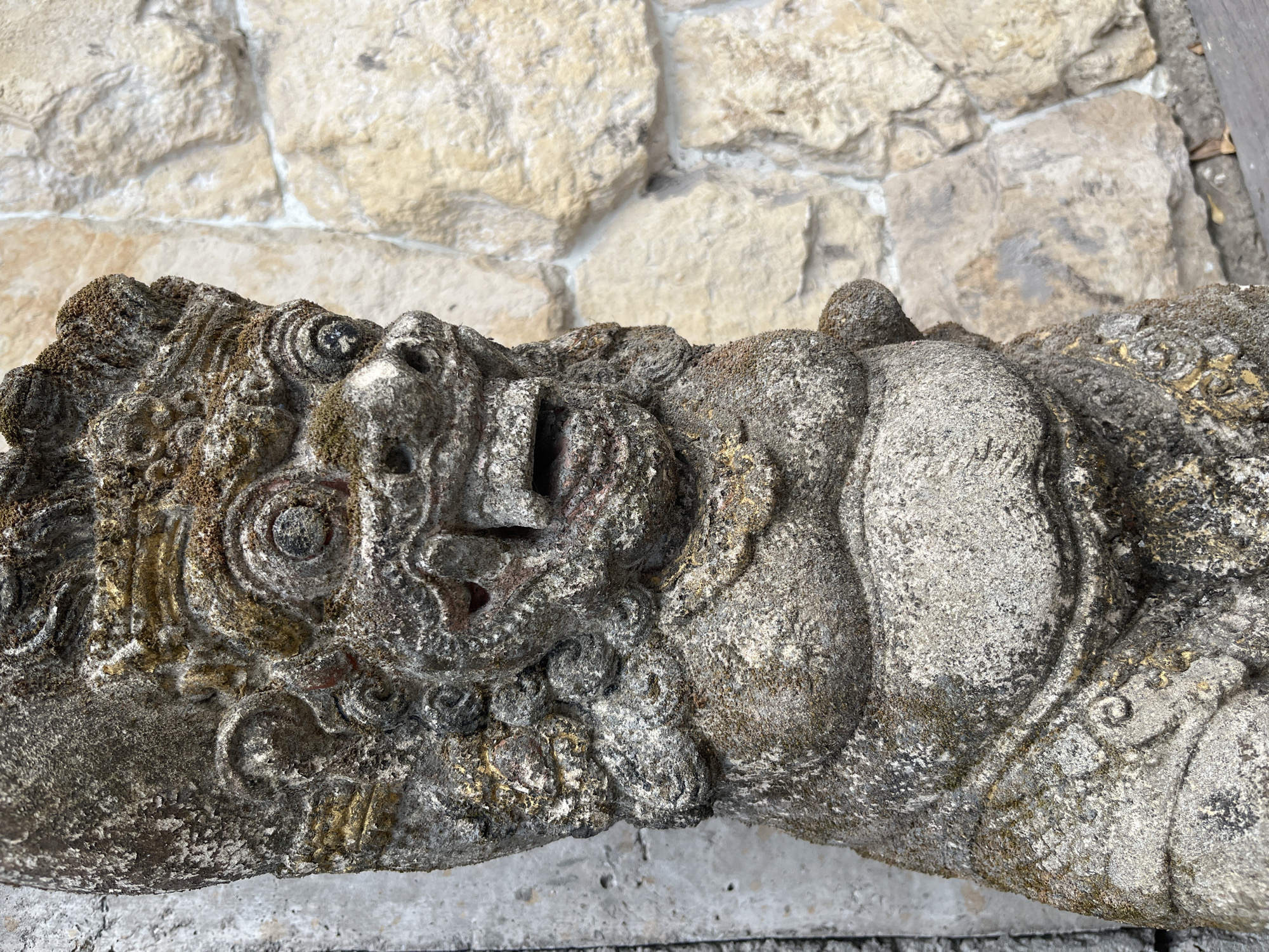 A weathered stone statue, resembling a mythical creature with a wide, open mouth and bulging eyes, stands in front of a stone wall. The statue is covered in patches of moss and appears to be ancient. Its body is bulky and adorned with intricate carvings, some of which retain remnants of gold paint. The background consists of a textured wall made of roughly cut, light-colored stones. The overall scene suggests an outdoor setting, likely a garden or temple in Kuta Selatan, Indonesia, given the style of the statue and the architecture.