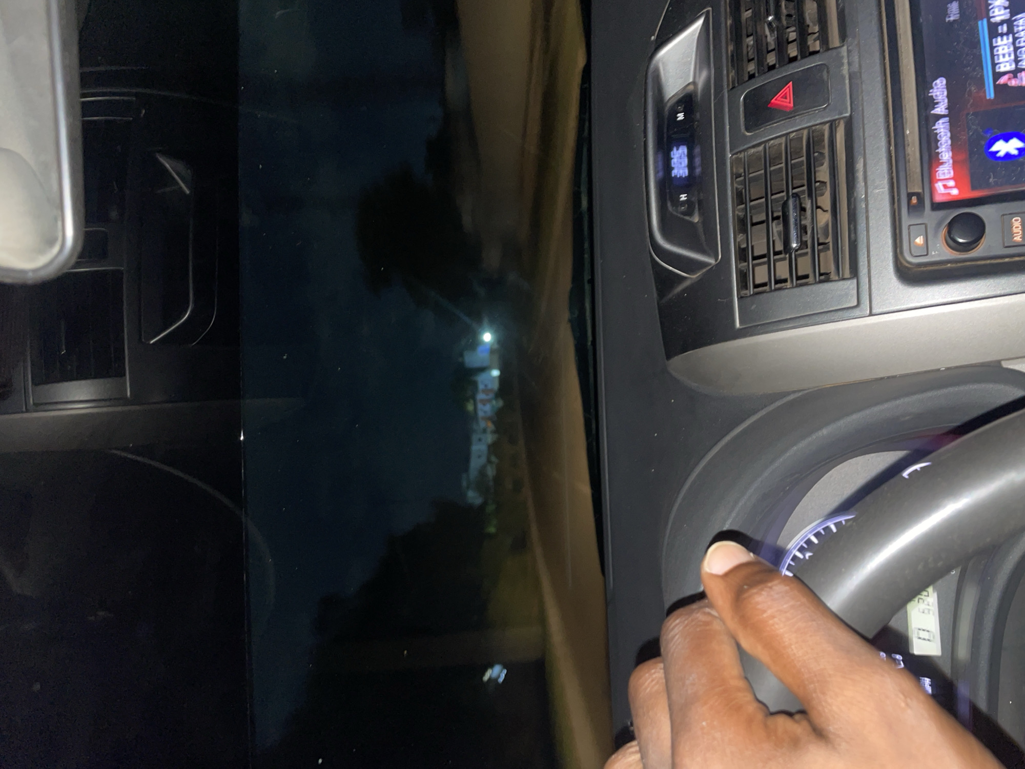 The image captures a sideways view from inside a car at night, focusing on the driver's side and the view through the windshield. A dark-skinned hand is visible on the steering wheel, suggesting a driver is present and likely operating the vehicle.
The car's interior features a dashboard with air vents, a prominent red triangular hazard light button, and a car stereo/infotainment system. The stereo display shows "BEBE - 1PM", indicating music or media, along with "Bluetooth Audio" and a Bluetooth icon, and "NO DATA". A digital display located to the left of the hazard button shows "5:05", likely the time, with letters "N" and "H" visible below it, possibly related to gear selection or driving modes. Part of the speedometer or tachometer is visible behind the hand on the steering wheel.
Through the windshield, a dark night sky is visible, punctuated by several bright, distant lights, which could be streetlights or lights from buildings. A faint outline of distant structures is also discernible. The time of day is clearly nighttime.
The scene depicts a journey or a stopped vehicle at night, within the context of Abuja, Nigeria.