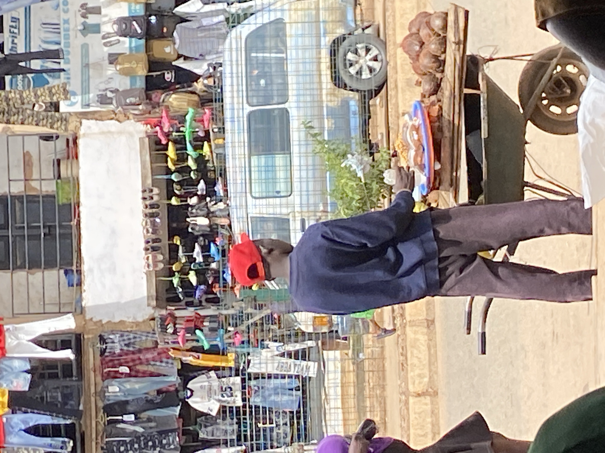 In Bukuru, Nigeria, a young man wearing a dark blue sweatshirt and a red baseball cap stands facing away from the camera, holding a small item in his right hand. He is positioned in front of a food cart laden with various items, including what appear to be coconuts and a platter of food. The cart itself is an old wheelbarrow with a wooden top. Behind him, a light-colored van with a checkered pattern is visible, partially obscured by a shrub. In the background, stalls display a variety of clothing and footwear, with some garments hanging from a clothesline. The scene depicts a bustling outdoor market environment.