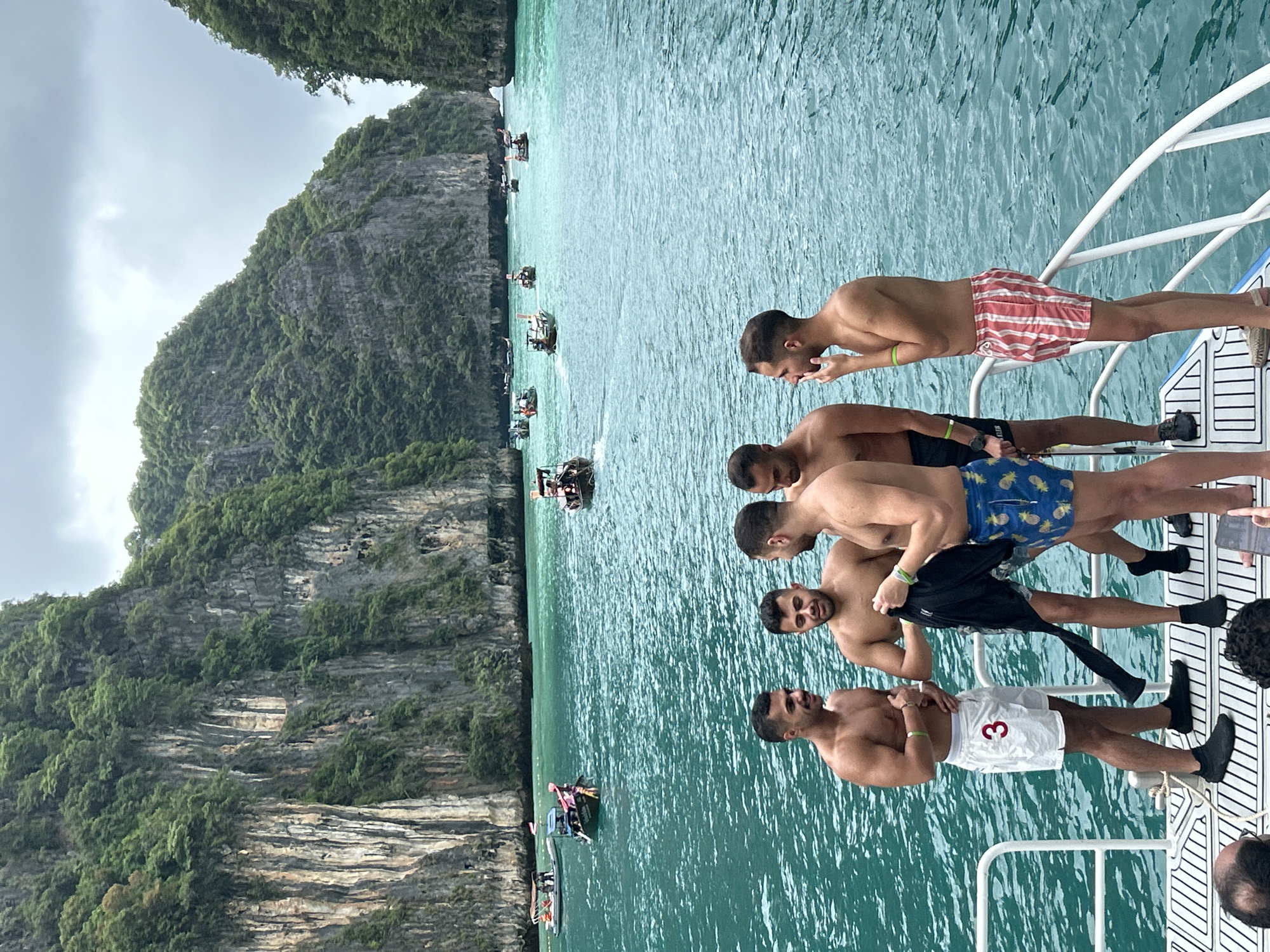 The image depicts a group of five young adult males on the deck of a boat, with a scenic backdrop of limestone karst formations and turquoise waters. The boat appears to be in Ao Nang, Thailand, a popular tourist destination known for its stunning coastal scenery and islands.
The five men are casually dressed, most in swimwear. From left to right, the first man is wearing white shorts with the number "3" in red. The second man is wearing dark swim trunks and holding a black garment. The third man is wearing blue swim trunks with a pineapple pattern. The fourth man is wearing red and white striped swim trunks. The fifth man's attire is not fully visible. They are engaged in conversation, with one man gesturing and another looking down.
The water is a vibrant turquoise, indicating clear and shallow conditions. Numerous longtail boats, a common mode of transport in this region, are scattered across the water, some carrying passengers and others appearing to be anchored. The limestone cliffs, characteristic of the Ao Nang region, rise dramatically from the water, covered in lush green vegetation. The sky is overcast with grey clouds, suggesting a diffused light that softens the scene. The overall atmosphere is relaxed and suggestive of a leisure outing or tour.