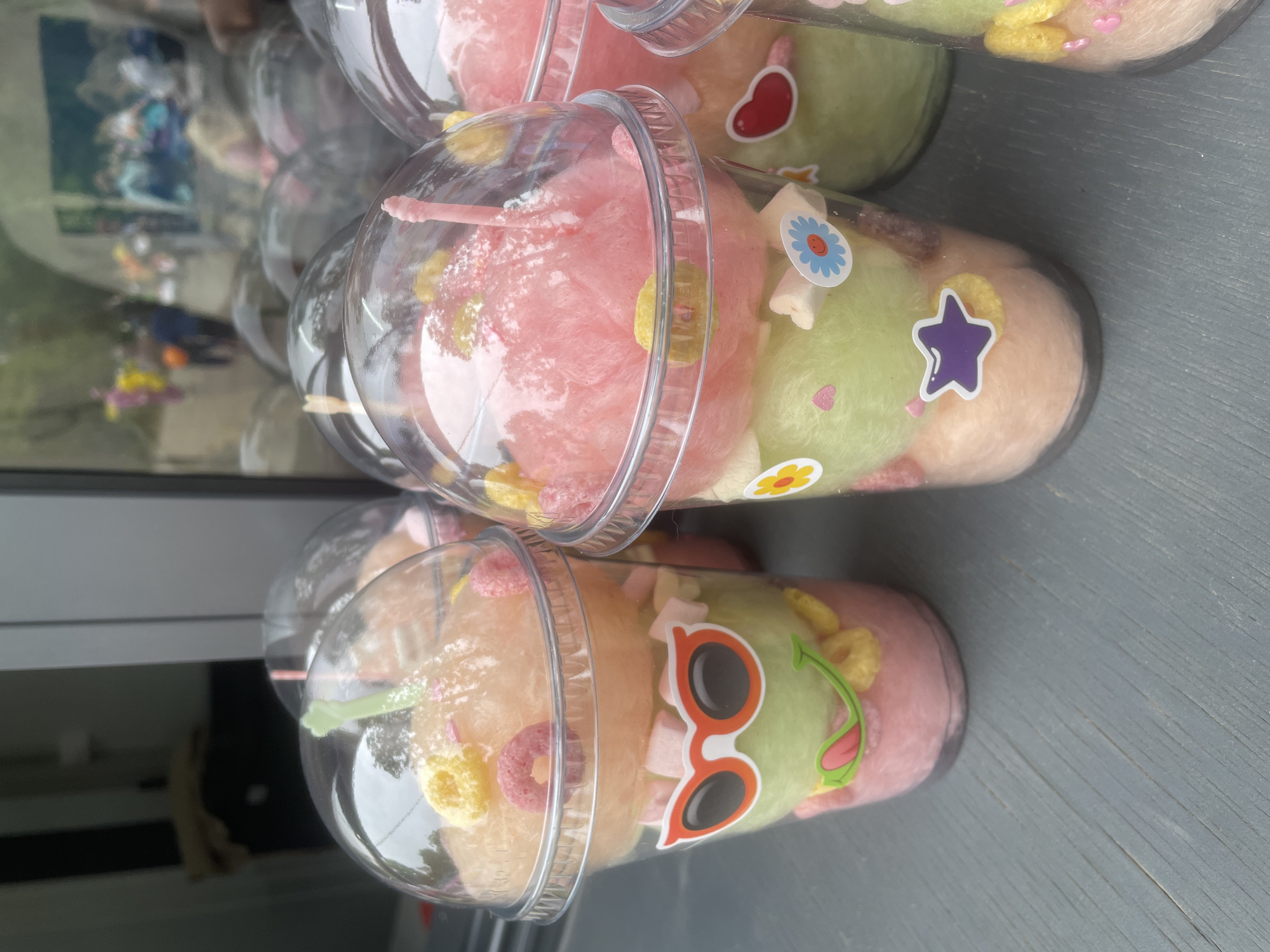 A display of multiple clear plastic cups with dome lids, containing layered, pastel-colored cotton candy, is positioned on a dark grey, horizontally oriented surface. The scene is illuminated by ambient light, suggesting a daytime setting. The cups contain distinct layers of pink, light green, and light orange cotton candy. Each cup is topped with various small candies or cereal pieces, including yellow crunchy bits, pink O-shaped cereal, and white star-shaped marshmallows. Several cups feature plastic straw-like implements, such as a light green one in the leftmost cup and a pink one in the central cup.
The cups are adorned with decorative stickers: The leftmost cup displays a frog-like face with orange sunglasses and a green smiling mouth with a protruding red tongue. The central cup features smaller stickers, including a blue flower, a purple star, and a yellow flower. A red heart sticker is visible on the rightmost partial cup.
The background of the image is dominated by a reflective surface, likely a window or glass, showcasing a blurred outdoor scene. This reflection reveals trees or foliage, indicative of a natural environment, possibly a park or public area. Multiple human figures are discernible in the reflection, some wearing brightly colored clothing (e.g., yellow, pink, blue), suggesting public activity. The precise actions and interactions of these reflected individuals are indistinct. This scene is located in Vladivostok, Russia.