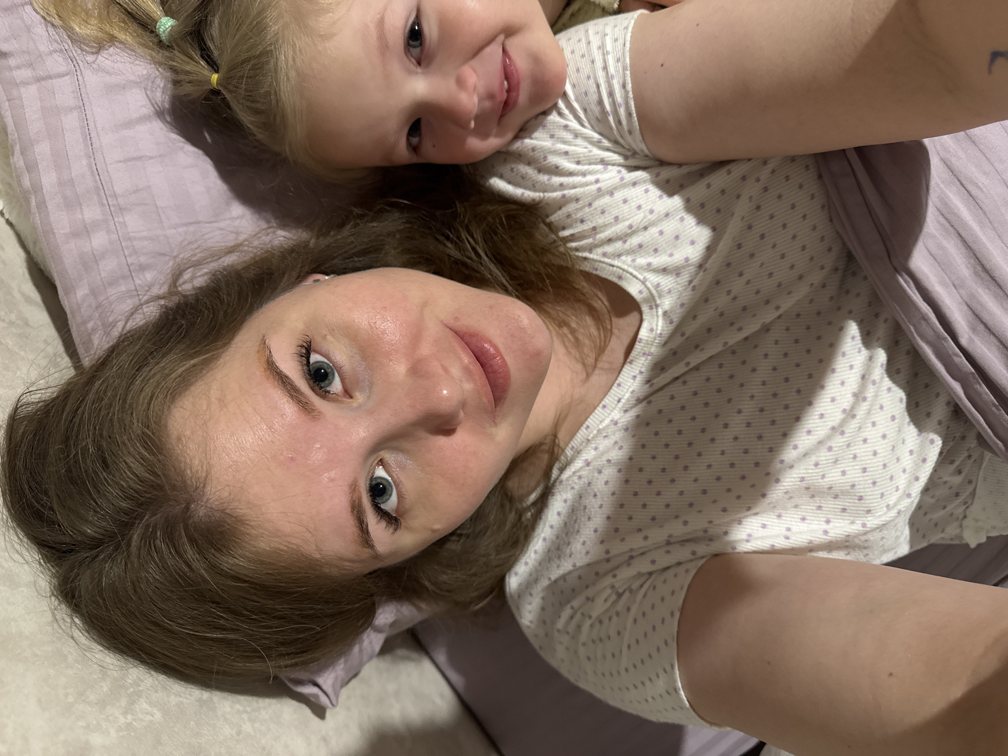 A high-angle selfie captures an adult and a young child lying down together in what appears to be a bed or sofa. The adult, positioned in the foreground, has light brown hair, blue eyes, and fair skin, looking directly at the camera with a gentle smile. She is wearing a white t-shirt adorned with small light purple polka dots.
Behind and slightly above the adult, a young child with blonde hair and blue eyes smiles brightly at the camera. Her hair is tied into two small pigtails, secured with light green and yellow elastic bands. She is also wearing a white top with light purple polka dots, similar to the adult's.
They are resting on light purple bedding, with a patterned pillow visible behind the child's head. The overall impression is one of warmth, affection, and happiness, suggesting a relaxed moment, possibly between a mother and child, in an indoor setting in Gornyi (Dvurechenskoe S/P), Russia. There is no visible text.