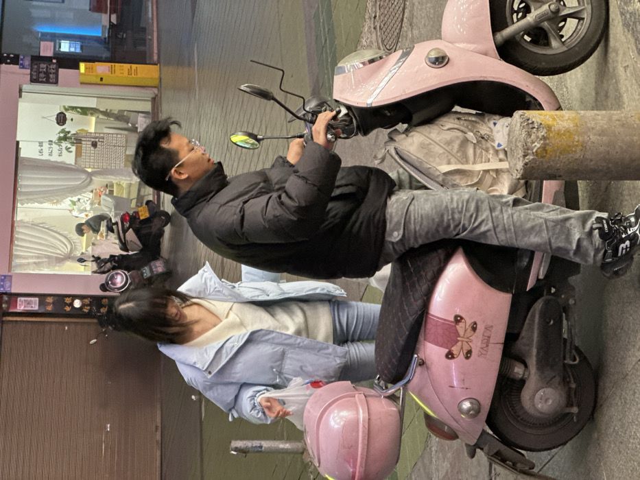 This image depicts an urban street scene in Jiangjin, China, at night, illuminated by artificial light. In the foreground, an adult male, wearing a dark puffy jacket, grey patterned trousers, and black sneakers, is seated on a pink scooter. He wears glasses and is looking down at the scooter's handlebars, with his hands near the controls. A light-colored backpack rests between his legs on the scooter's footrest. To the left of the man and scooter, an adult female stands, wearing a light blue puffy jacket, a cream V-neck sweater, and light blue jeans. She holds a clear plastic bag with contents in her left hand, looking downwards towards the bag or scooter. The pink scooter features a black textured seat, a pink storage box on the rear rack, and a brown butterfly decal with the word "YANJIA" on its side panel. A grey concrete bollard with yellow markings partially obscures the front wheel of the scooter. In the background, across a street or pedestrian path, multiple shopfronts are visible with interior lighting. One brightly lit shop features white curtains and a seated individual inside. Another scooter is parked further back. To the right, a yellow outdoor payment kiosk, labeled "ALIPAY" and with Chinese characters, stands next to a building wall.