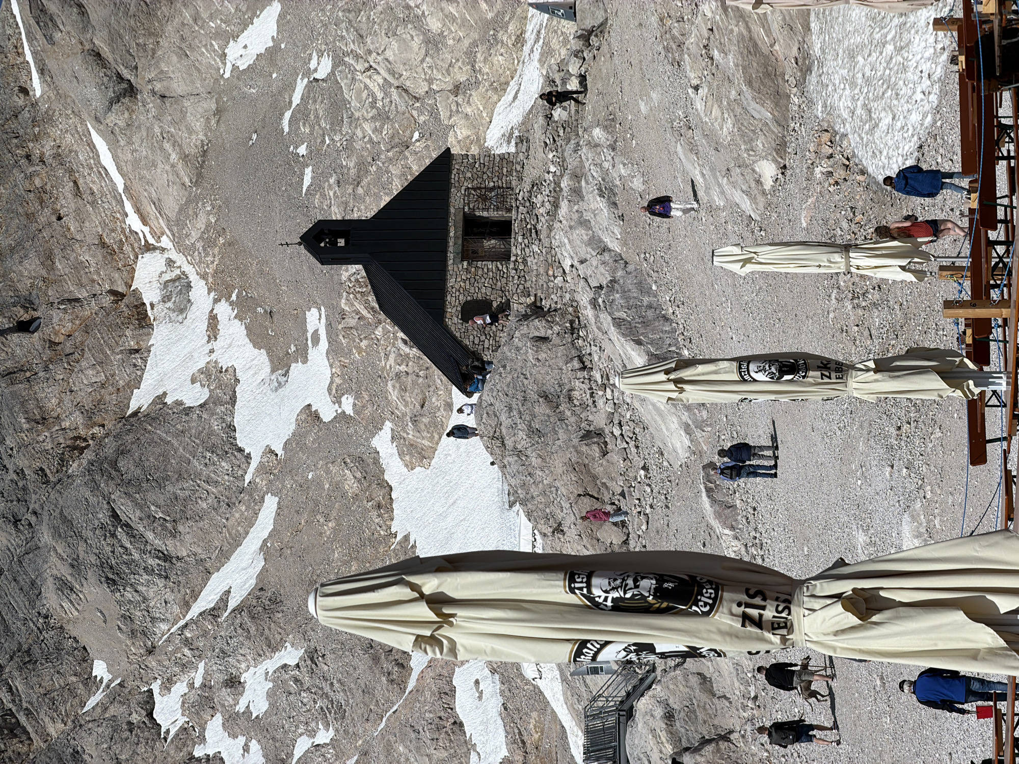 The image depicts a mountainous landscape with sparse vegetation and patches of snow, characteristic of a high-altitude environment. In the mid-ground, a small stone building with a black, pitched roof and a bell tower stands on a rocky outcrop. Several individuals are scattered across the rocky terrain, some walking and others gathered near the building. In the foreground, several large, cream-colored umbrellas are positioned, some of which are closed and appear to be part of an outdoor seating area, possibly a restaurant or cafe. The context suggests this location is likely near the summit of a mountain accessible to tourists, given the presence of the building, the umbrellas, and the people. This scene is consistent with locations found in the Zugspitze massif near Garmisch-Partenkirchen, Germany, a popular tourist destination offering hiking and panoramic views.