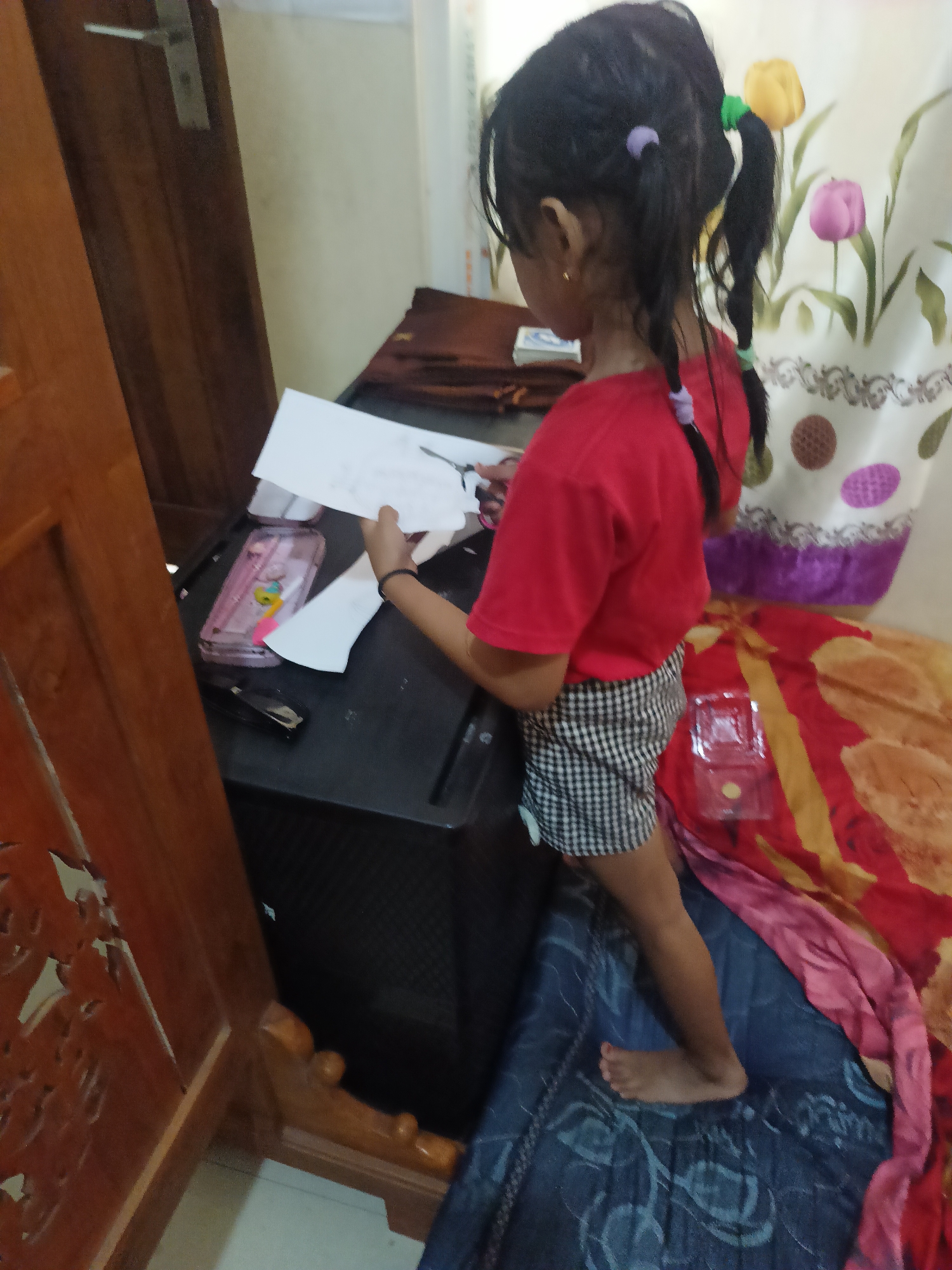 The image depicts a young girl, likely of Indonesian ethnicity, standing on a bed and leaning over a dark-colored surface, possibly a table or desk. She is wearing a red t-shirt and a black and white checkered skirt. Her dark hair is styled in pigtails with purple and green hair ties. In her hands, she holds a piece of white paper and appears to be using scissors, suggesting she is engaged in a craft or cutting activity. To her left, on the dark surface, a pink pencil case with colorful designs and a black stapler are visible. In the background, a curtain with a floral pattern featuring tulips in pink and yellow, along with some circular designs, is present. The bed she is standing on is covered with a brightly colored bedspread in shades of red, yellow, and pink. The environment appears to be an indoor residential setting. Based on the provided context, this scene is situated in Rengging, Indonesia.
