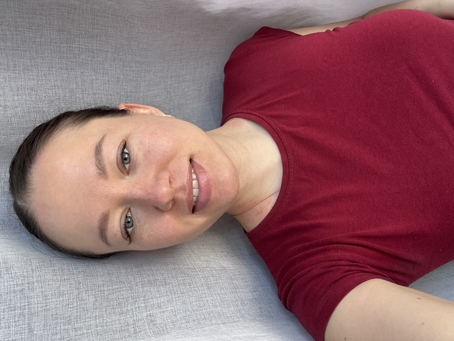 This image depicts a single adult female in an indoor setting within the city of Shul'gino, Russia. The subject, estimated to be between 25 and 35 years old, has fair skin with scattered freckles visible on her forehead, nose, and cheeks. Her dark hair is pulled back, revealing a clear hairline. She possesses greenish-blue eyes with enhanced eyelashes and well-defined eyebrows. Her lips have a glossy appearance, and she displays a closed-mouth smile, revealing her upper teeth. Small, silver-toned stud earrings are present in both earlobes. Erythema, consistent with skin irritation or a rash, is visible on her lower neck and upper chest area. She is wearing a solid dark red, short-sleeved, scoop-neck t-shirt. A thin, gold-toned chain necklace is partially visible around her neck. The subject is positioned directly facing the viewer, looking into the camera lens, indicative of a posed photograph or selfie, with her left upper arm partially visible in the lower-left corner of the frame. The background consists of a uniformly lit, light-colored (likely off-white or light grey) textured fabric, consistent with a curtain or drapery. No other objects or details are discernible in the immediate environment.