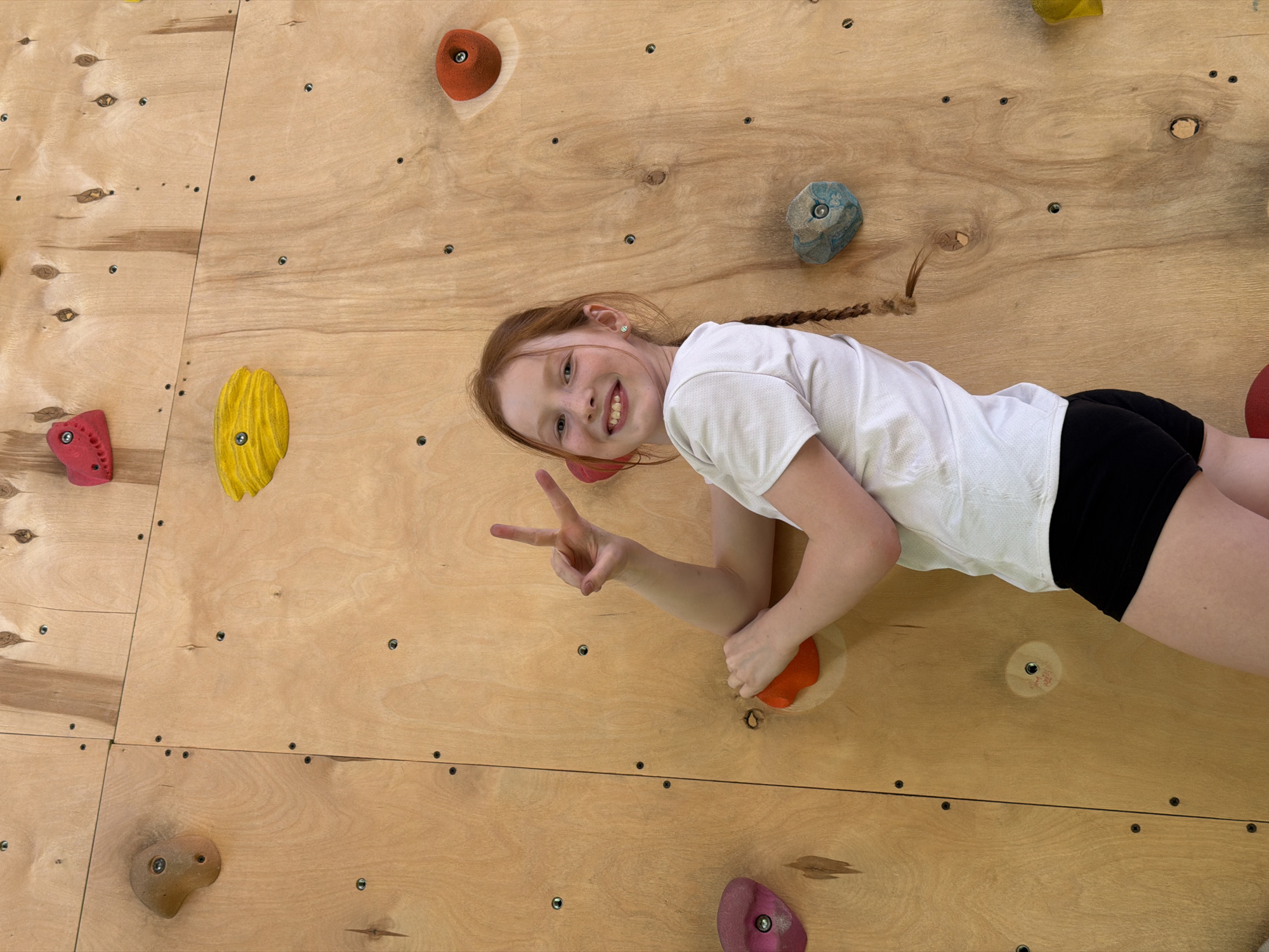 A young red-haired girl with a braid smiles and holds up a peace sign while positioned on a wooden climbing wall with colorful handholds. She is wearing a white t-shirt and black shorts. The wall consists of multiple sections of plywood joined together. The scene likely takes place in an indoor climbing gym; the location in Izhevsk, Russia is not visually discernible.
