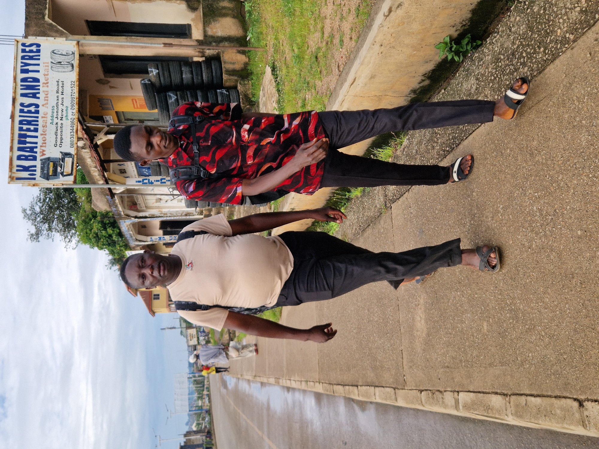 Two men walk side-by-side on a concrete sidewalk next to a wet asphalt road. The man on the left wears a tan t-shirt and dark pants. He has a backpack on his shoulders. The man on the right wears a red and black patterned shirt and dark pants. He also wears a backpack. Behind them, a sign reads "I.K BATTERIES AND TYRES WHOLESALE AND RETAIL," with an address listed below. Several tires are stacked to the right of the building. In the background, other pedestrians and vehicles are visible on the road, and buildings line the street. The sky is overcast. The scene appears to be located on Jonathan Road, opposite New Jos Hotel, in Jos, Nigeria.