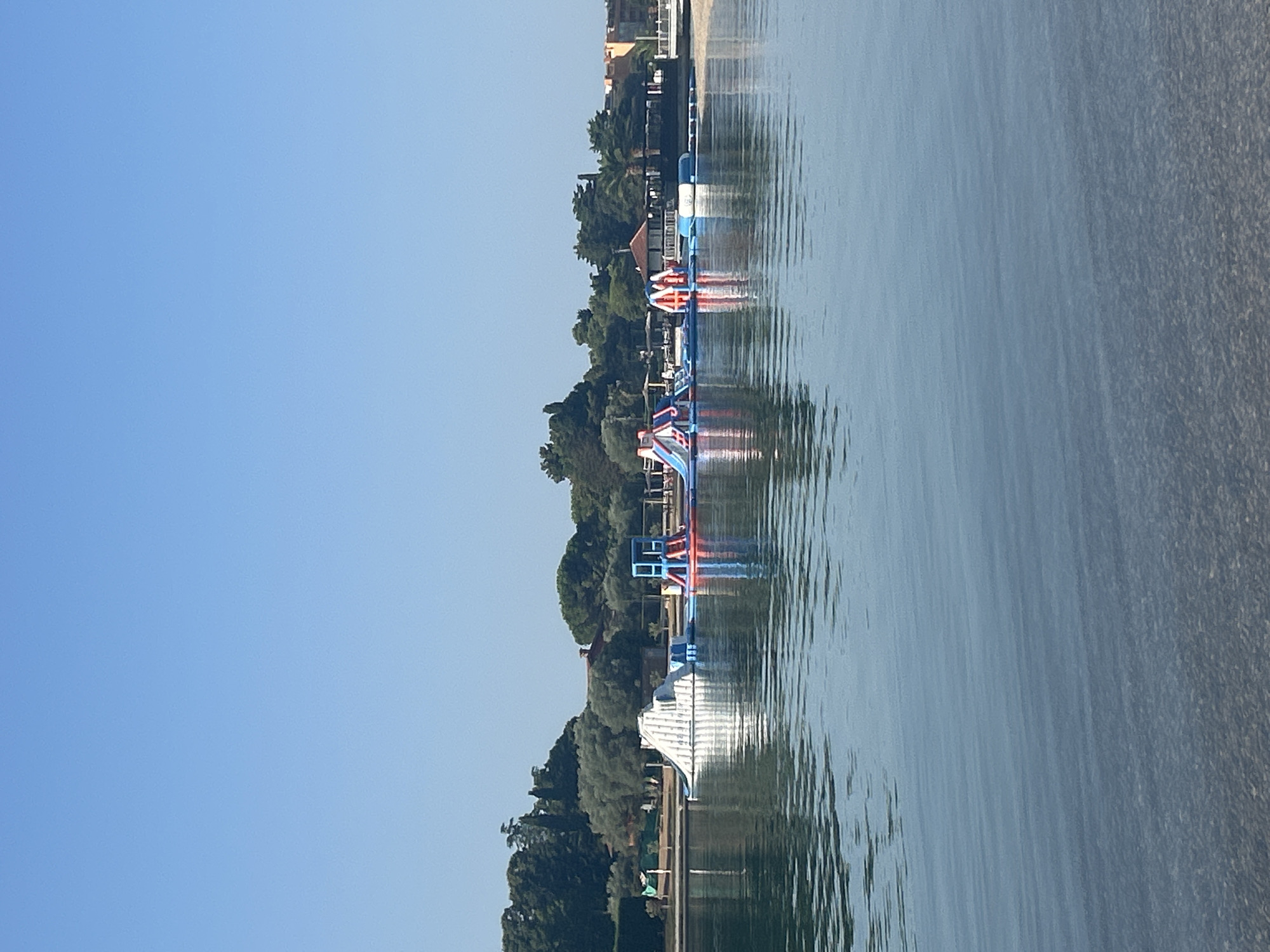 The image shows a clear blue sky over a body of water with a beach and trees in the background. In the water, there is an inflatable water park with various slides and climbing structures. The water park is located in front of a shoreline with vegetation and buildings. The water is calm and reflective, showing the colors of the sky and the water park. The scene appears to be a recreational area, possibly a beach resort, in Umag, Croatia. There are no people visible in the image. The overall impression is one of a peaceful summer day with a focus on water-based activities.
