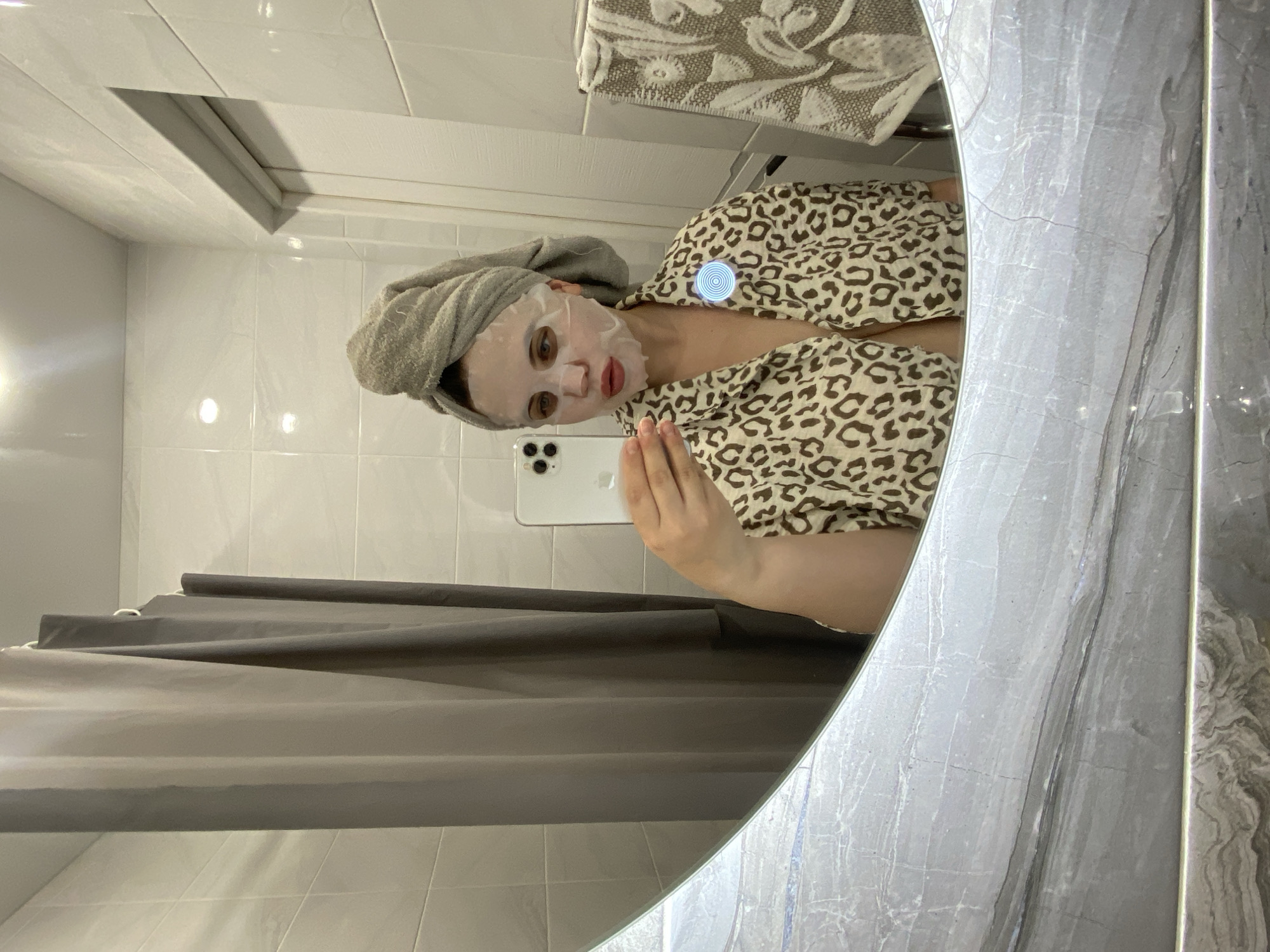 A person is taking a selfie in a bathroom. The reflection in a round mirror reveals a woman wearing a facial mask and a towel on her head, clad in a leopard-print robe. She is holding a smartphone. A gray shower curtain hangs behind her, and white tiles cover the walls. A towel is also hanging above. This is likely taken in the city of Chernushka, Russia.