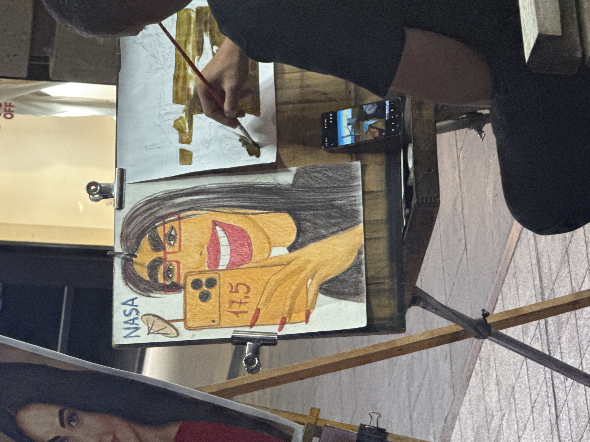 An overhead view captures an artist focused on painting, likely indoors at an art stand or studio. The artist, seen from the head, right arm, and torso, wears a dark shirt and holds a red-handled paintbrush, applying brown paint or ink to a paper secured by clips. This paper displays a light brown sketch, with outlines of figures or objects that are difficult to discern clearly.
Prominently displayed in the foreground is a vibrant caricature drawing of a woman with dark hair, red-framed glasses, and a wide, open-mouthed smile revealing white teeth. She is depicted holding a gold-colored smartphone, which has three camera lenses, as if taking a selfie. Her hand has long, red fingernails. Text "NASA" is written vertically on the left side of this drawing, and "17.5" is visible on the phone screen within the caricature.
To the artist's right, a smartphone is propped upright, displaying an image that appears to be a landscape or a reference photograph, though details are indistinct. Another drawing, possibly a portrait featuring a woman in red, is partially visible at the bottom left of the frame. In the background, a white sign shows a partial word, "OFF". The overall scene depicts an artistic process in progress, with finished work on display.