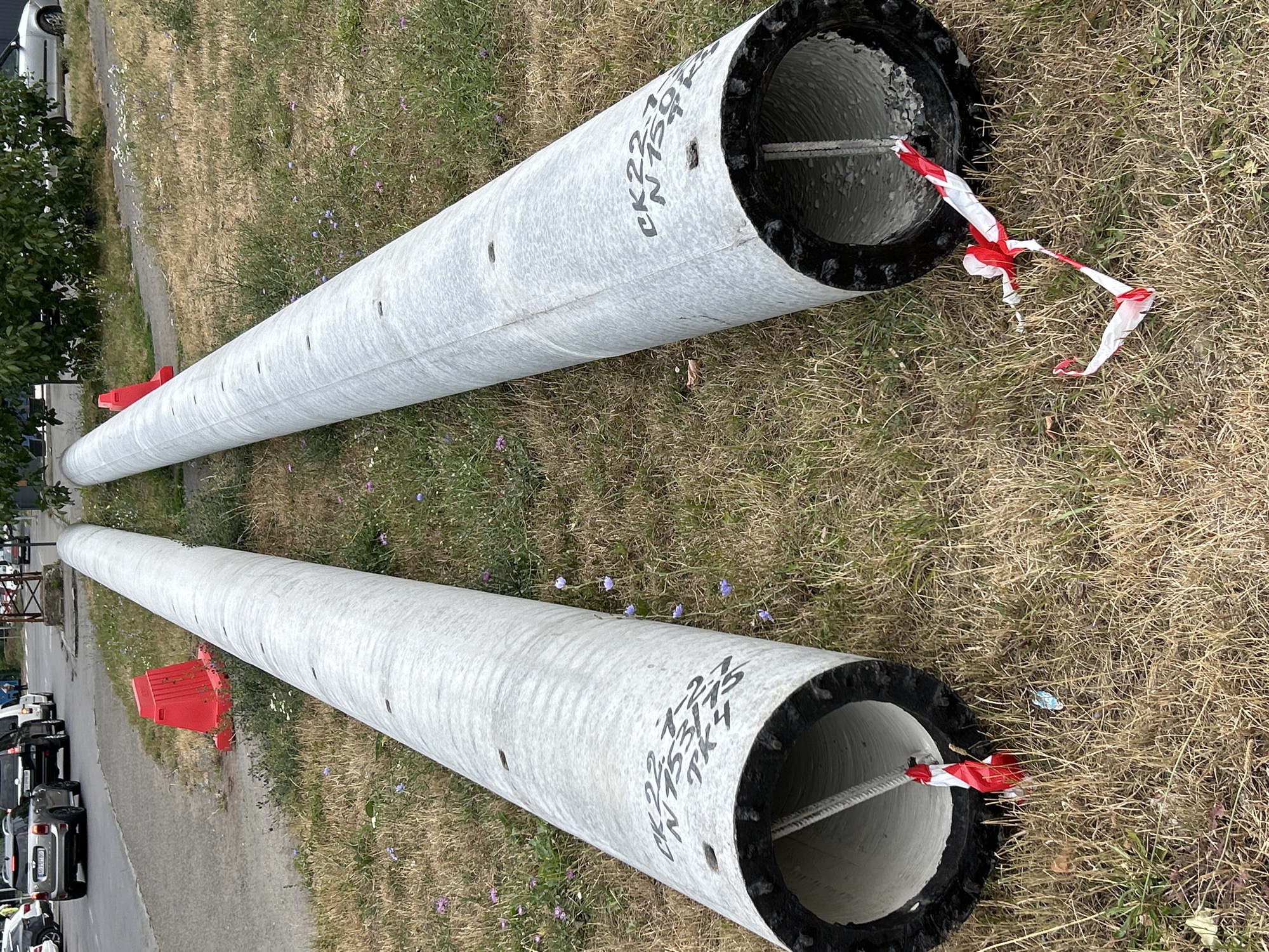 Two large, grey concrete pipes are lying horizontally on a patch of dry grass with scattered purple and white wildflowers. The pipes are parallel to each other, with the left pipe closer to the viewer. Both pipes have black, ribbed rims at their open ends. Red and white striped caution tape is tied to a rope inside the left pipe and trails out onto the grass. The right pipe also has caution tape tied to its internal rope. Black lettering is visible on the surface of both pipes. In the background, a paved road with vehicles is visible, including a silver Mini Cooper, a black SUV, and a white van. A red traffic cone is situated on the grass between the road and the pipes. Trees and foliage are present further in the background. The scene appears to be a roadside location within the city of Uzhhorod, Ukraine.