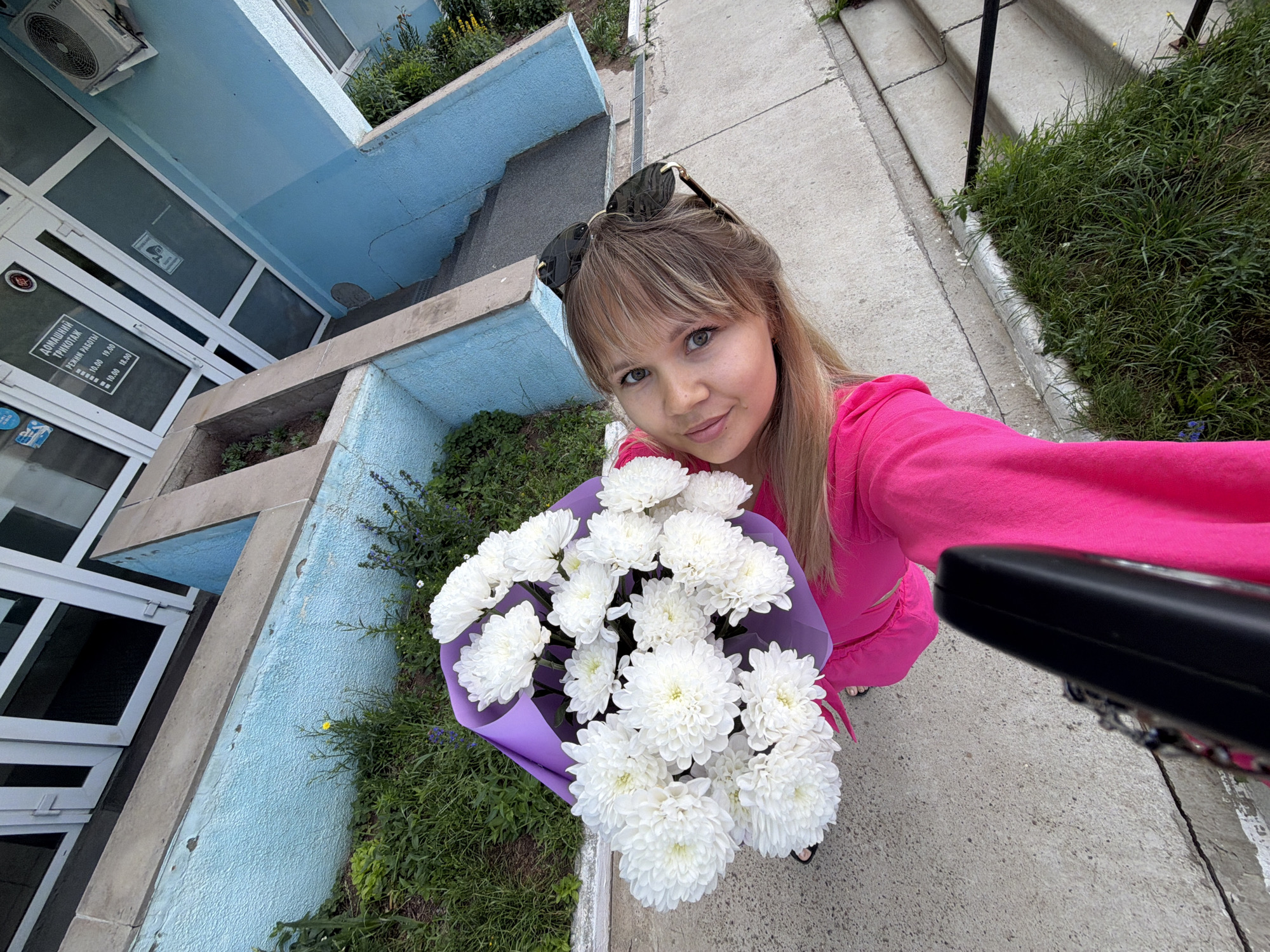 A young woman with blonde hair and blue eyes holds a bouquet of white chrysanthemums wrapped in purple paper. She is wearing a bright pink hooded jacket and sunglasses are perched on her head. The woman is standing on a concrete walkway next to a grassy area and a blue painted building with a set of stairs leading down. A black object, possibly a camera or phone, is held by the woman, angled towards herself for a selfie. A sign with Cyrillic text is visible on the building. The scene appears to be outdoors in an urban environment, likely the city of Chaikovsky, Russia, given the building's signage.
