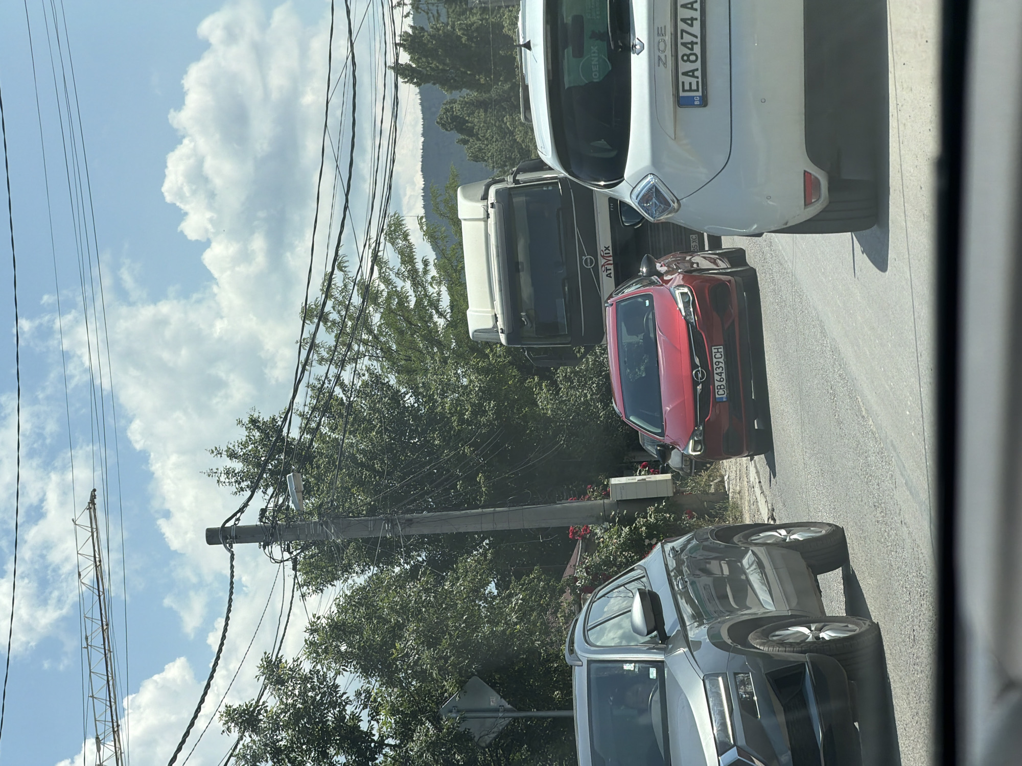 The image shows a traffic scene with several cars on a road. In the foreground, the rear of a white electric car, identified by the "ZOE" badge and license plate "EA 8474 A BG," is visible. To its left is a red Opel Astra with license plate "CB 6439 CH." Further left, a grey SUV, possibly a Skoda, is partially visible. In the background, a large white truck with "AT MIX" branding is stopped in traffic. The environment appears to be a suburban or semi-urban area with trees lining the road and power lines overhead. In the distance, a mountainous landscape can be seen. The sky is blue with scattered clouds. The scene suggests a standstill or slow-moving traffic situation. Based on the license plate format "EA 8474 A BG" and the road infrastructure, the location is identified as Sofia, Bulgaria.