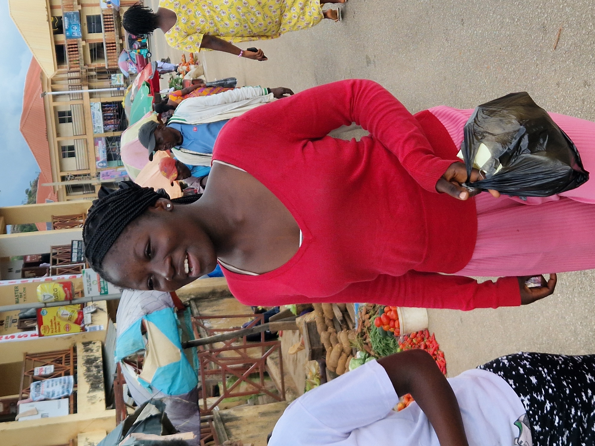 A medium shot captures a young Black woman with braided hair, wearing a red long-sleeved shirt and a pink skirt, holding a black plastic bag in her right hand while walking forward. Her left arm is bent at the elbow, and she is looking to her left with her mouth slightly open. In the background, a market scene unfolds. Various stalls are visible, with vendors selling produce like tomatoes and other vegetables. Umbrellas are used to provide shade. Buildings with shops line the street, and people are seen walking around, some wearing casual attire. The overall setting appears to be a bustling outdoor market in a Nigerian city.