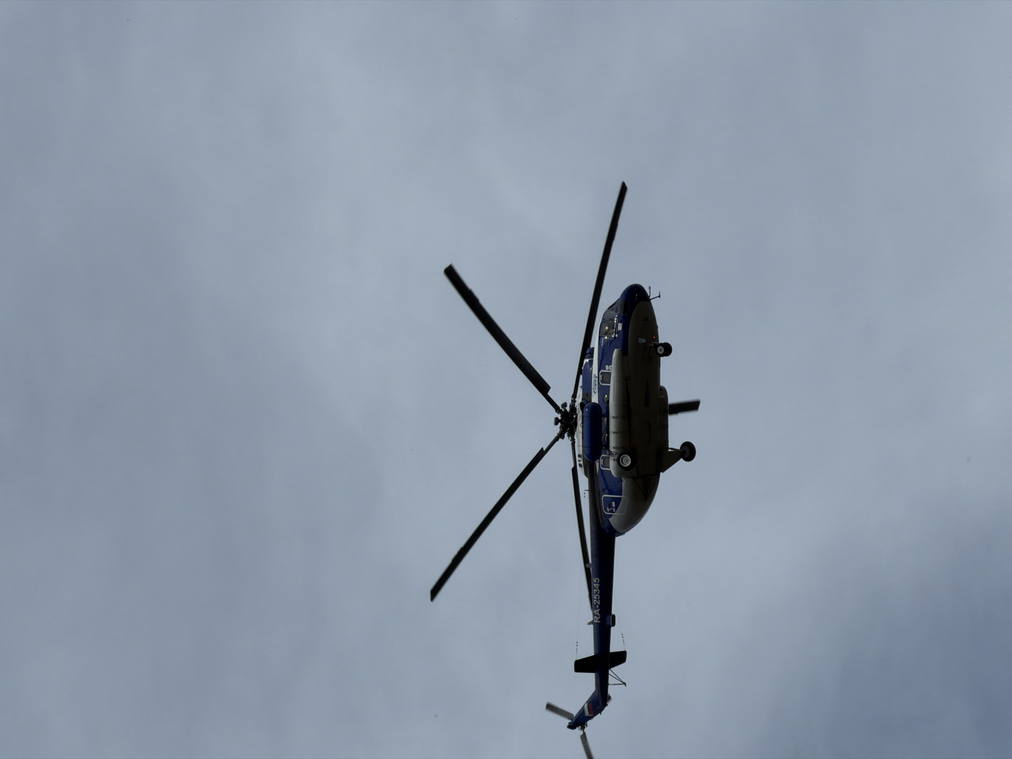 A blue and white helicopter with the registration "RA-25345" is shown in flight against a cloudy sky. The helicopter has a prominent rotor system with four blades. The scene appears to be an aerial view, and while the location is specified as Chaikovsky, Russia, there are no visible landmarks or contextual elements within the image to confirm this. The sky is uniformly overcast, with no clear indicators of time of day or specific weather conditions beyond cloud cover. There are no people or other objects clearly visible in the frame.
