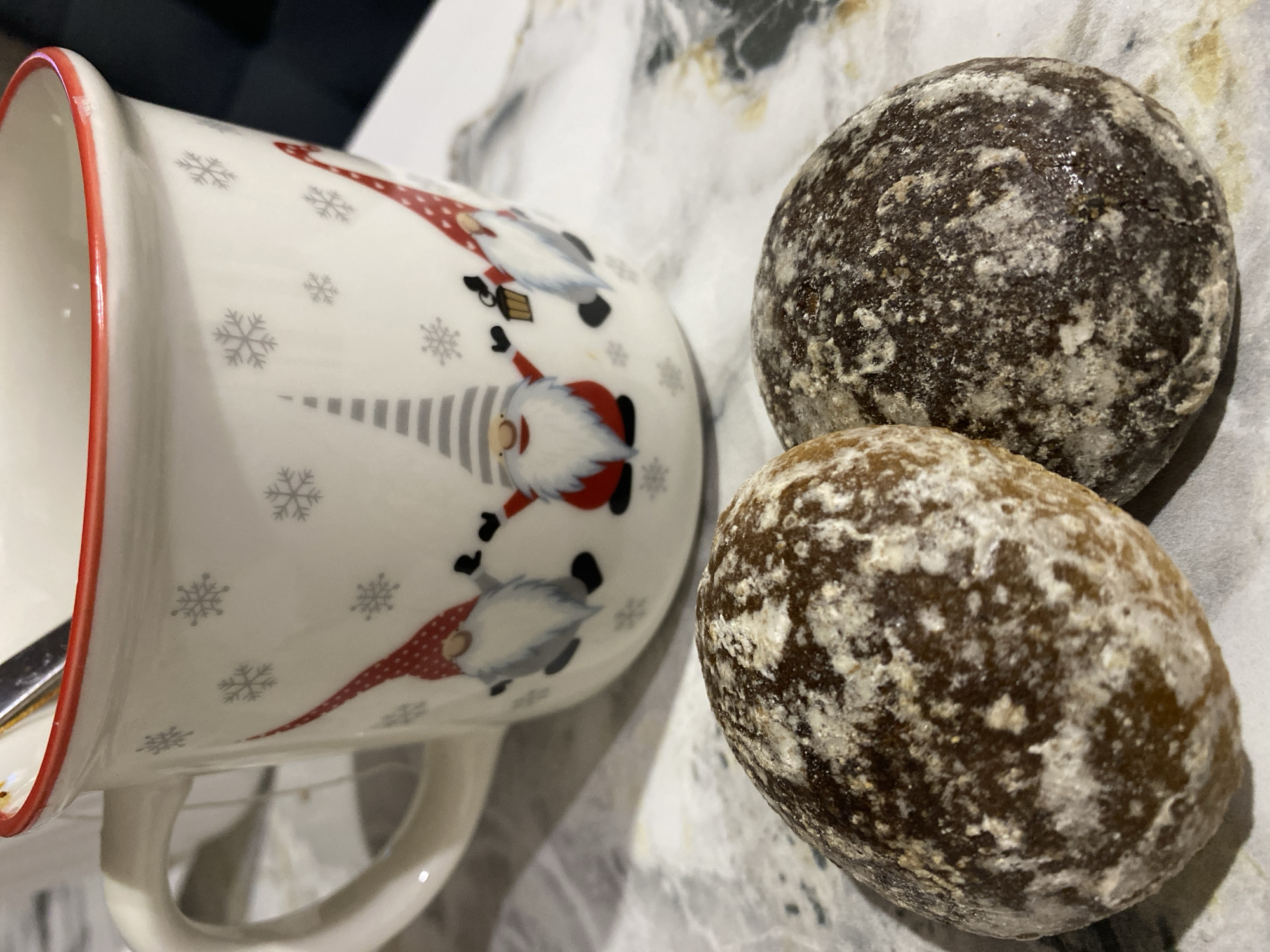 An indoor scene in the city of Chaikovsky, Russia, depicts a white ceramic mug with a red rim and handle interior, decorated with grey snowflakes and three gnomes wearing red or striped conical hats. A silver spoon is partially visible inside the mug, which contains an unidentifiable liquid. Positioned in front of the mug are two round, dark brown cookies, both partially covered with an uneven dusting of white powder. The items rest on a light-colored surface featuring a grey, marble-like veining pattern.