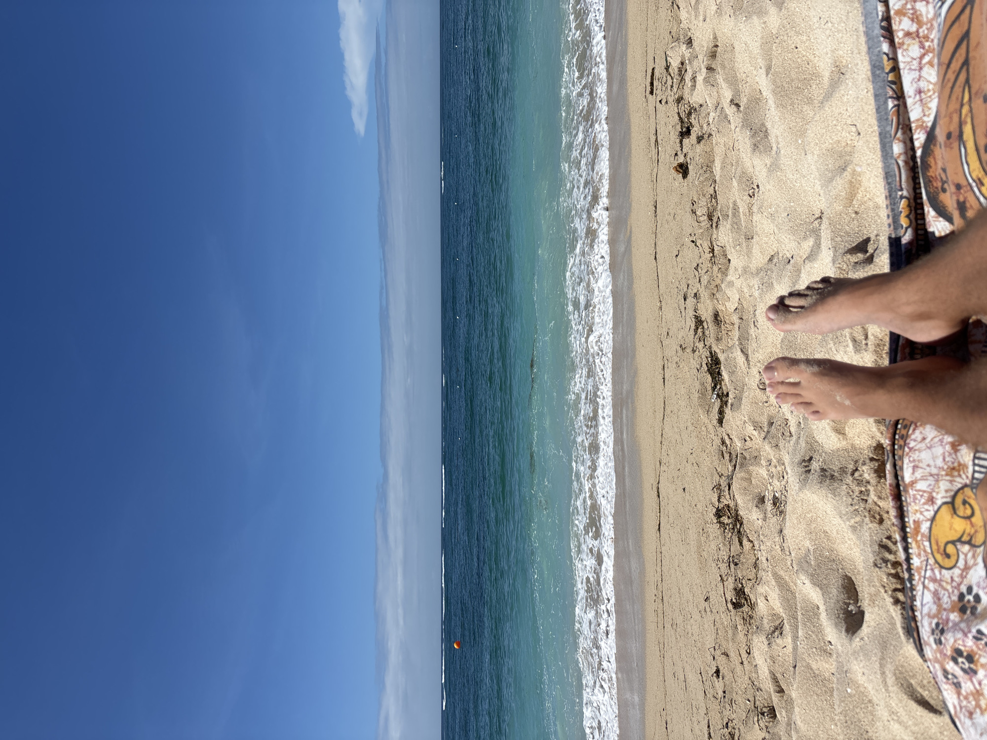 The image shows a view of a beach in Kuta Selatan, Indonesia. The perspective is from someone lying on a patterned sarong on the sand, with their feet visible. The sand is light brown, and there is seaweed scattered on it. Beyond the sand, the turquoise ocean meets the shore with foamy white waves. In the distance, the ocean transitions to a darker blue. The sky above is clear blue with a few wispy white clouds near the horizon. An orange buoy is visible in the water.