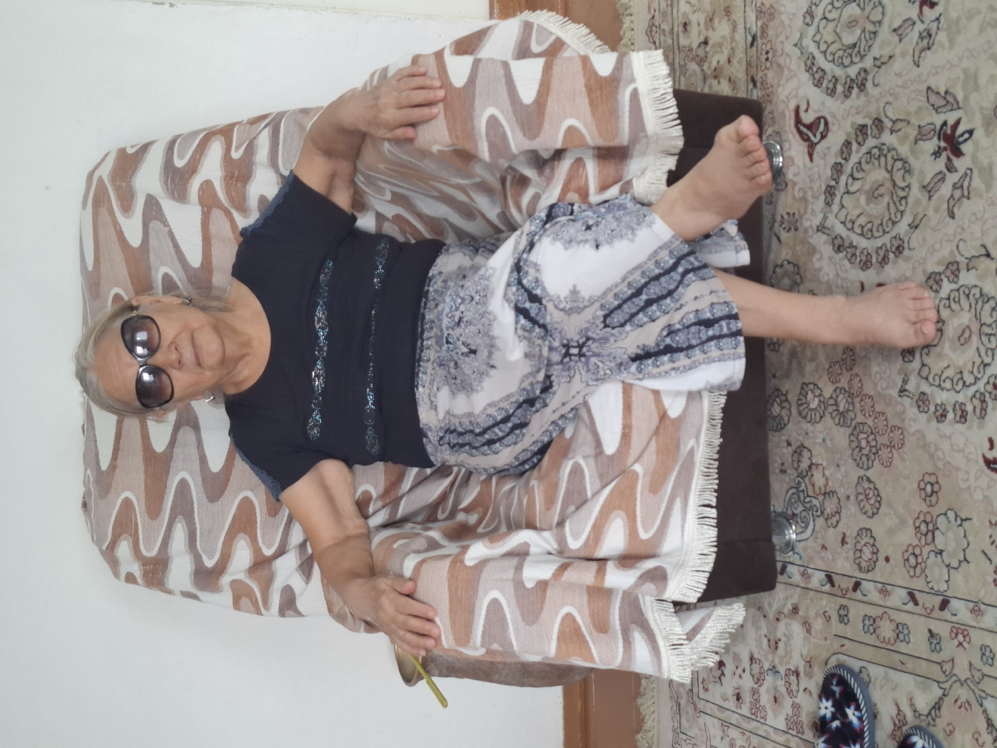 A full shot shows an elderly woman with gray hair wearing sunglasses and a dark shirt with a patterned skirt, seated in a large armchair. The armchair is draped with a brown and white wavy patterned blanket. The woman is barefoot and her legs are crossed. She is in a room with a white wall and a patterned rug on the floor. On the left side of the image, there are a pair of slippers. The image appears to be taken in an indoor domestic setting.