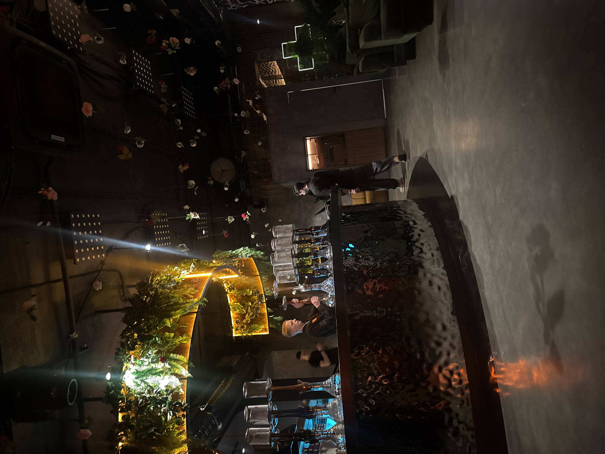 The image depicts the interior of a dimly lit hookah lounge. The scene is dominated by a dark, textured bar in the foreground, reflecting ambient orange and yellow lights. Behind the bar, several hookah pipes are displayed on shelves, illuminated by subtle lighting. A man with blond hair, wearing a black shirt and an orange insignia, is standing at the bar, appearing to be an employee. Another person in a dark shirt is visible further back. Overhead, the ceiling features exposed pipes and is adorned with artificial greenery and scattered faux flowers. The background reveals lounge seating areas with dark green upholstery and a large neon cross-shaped installation. The flooring is a polished concrete surface.