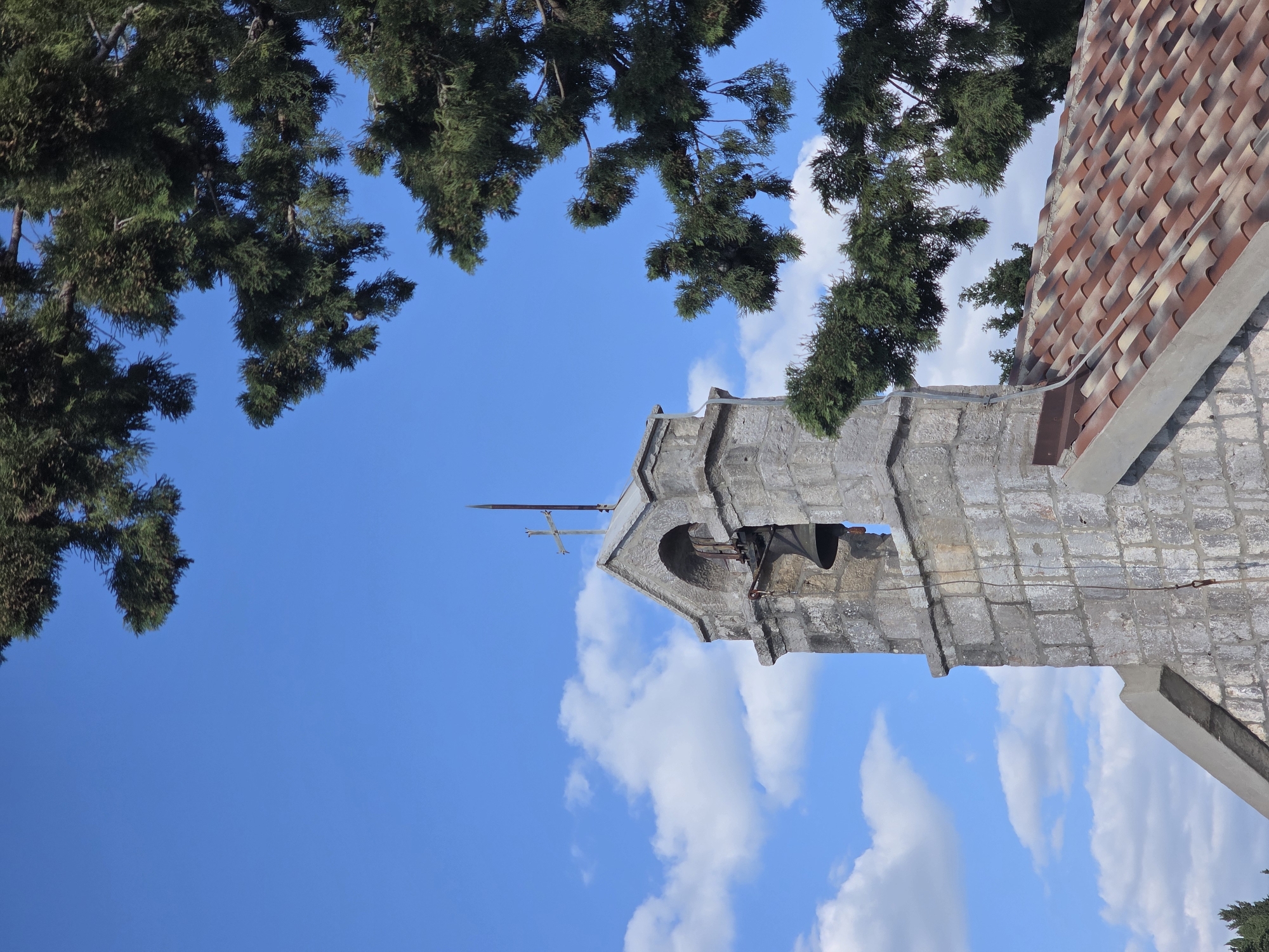 A low-angle shot depicts a stone bell tower with a red-tiled roof, set against a backdrop of a bright blue sky dotted with fluffy white clouds and framed by the green foliage of tree branches. The tower is made of weathered gray stone blocks and features an arched opening in which a bell hangs. On top of the tower is a cross and a lightning rod. The scene is bathed in sunlight, suggesting a clear day. The location is Luštica, Montenegro.