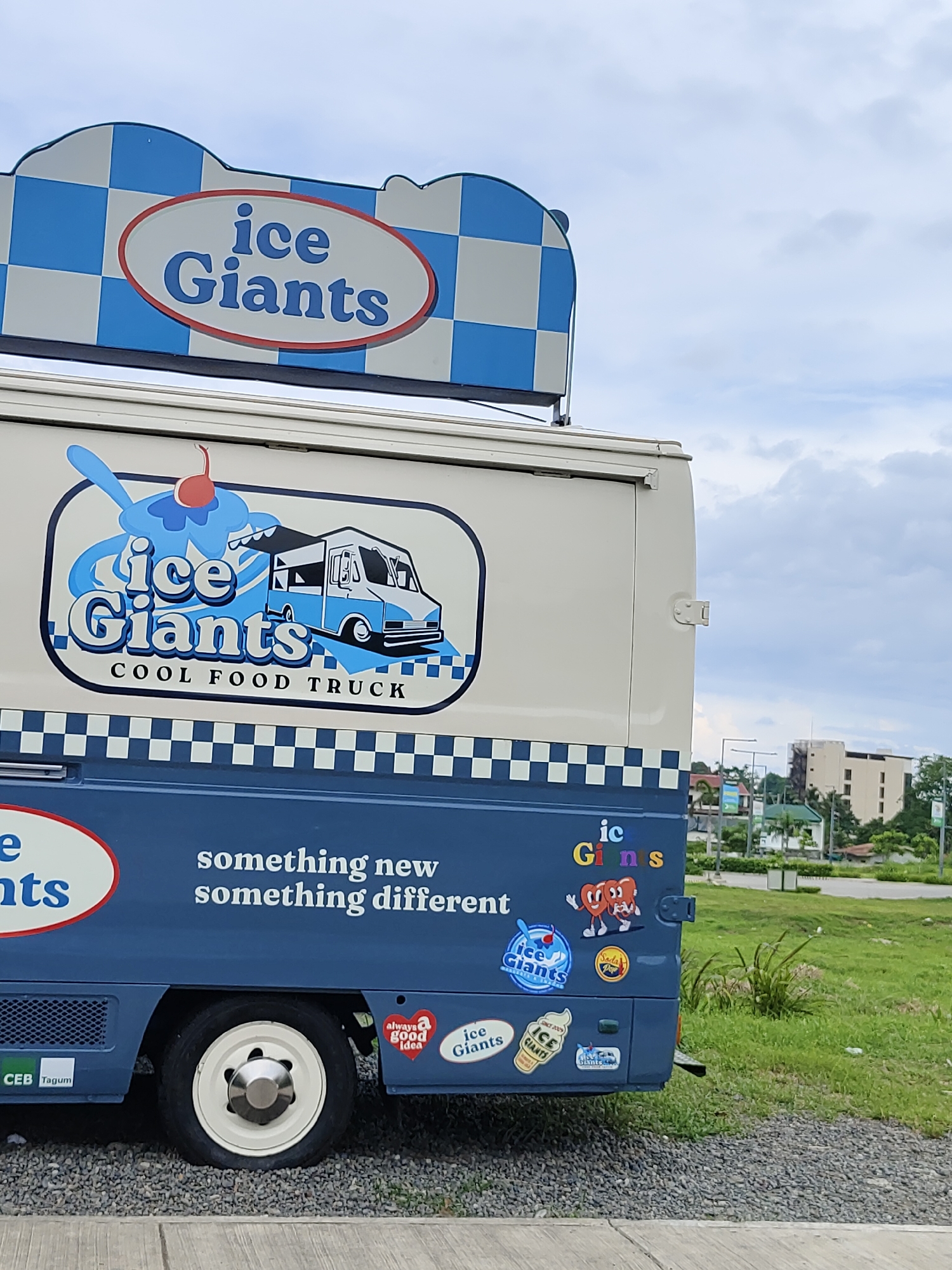 A side view of a blue and cream-colored "Ice Giants" food truck is visible, parked on a gravel surface. The truck features branding with the "Ice Giants" logo, including a stylized ice cream cone and a blue and white checkered design. The slogan "something new something different" is also displayed on the side of the truck in white lettering. Several stickers are affixed to the truck, including one with two heart characters, a "Seda Pop" sticker, and various "Ice Giants" branded stickers. In the background, a building with a light-colored facade and trees can be seen, suggesting an urban or suburban setting. The sky is overcast with clouds. A white wheel with a chrome hubcap is prominent on the lower left of the truck.