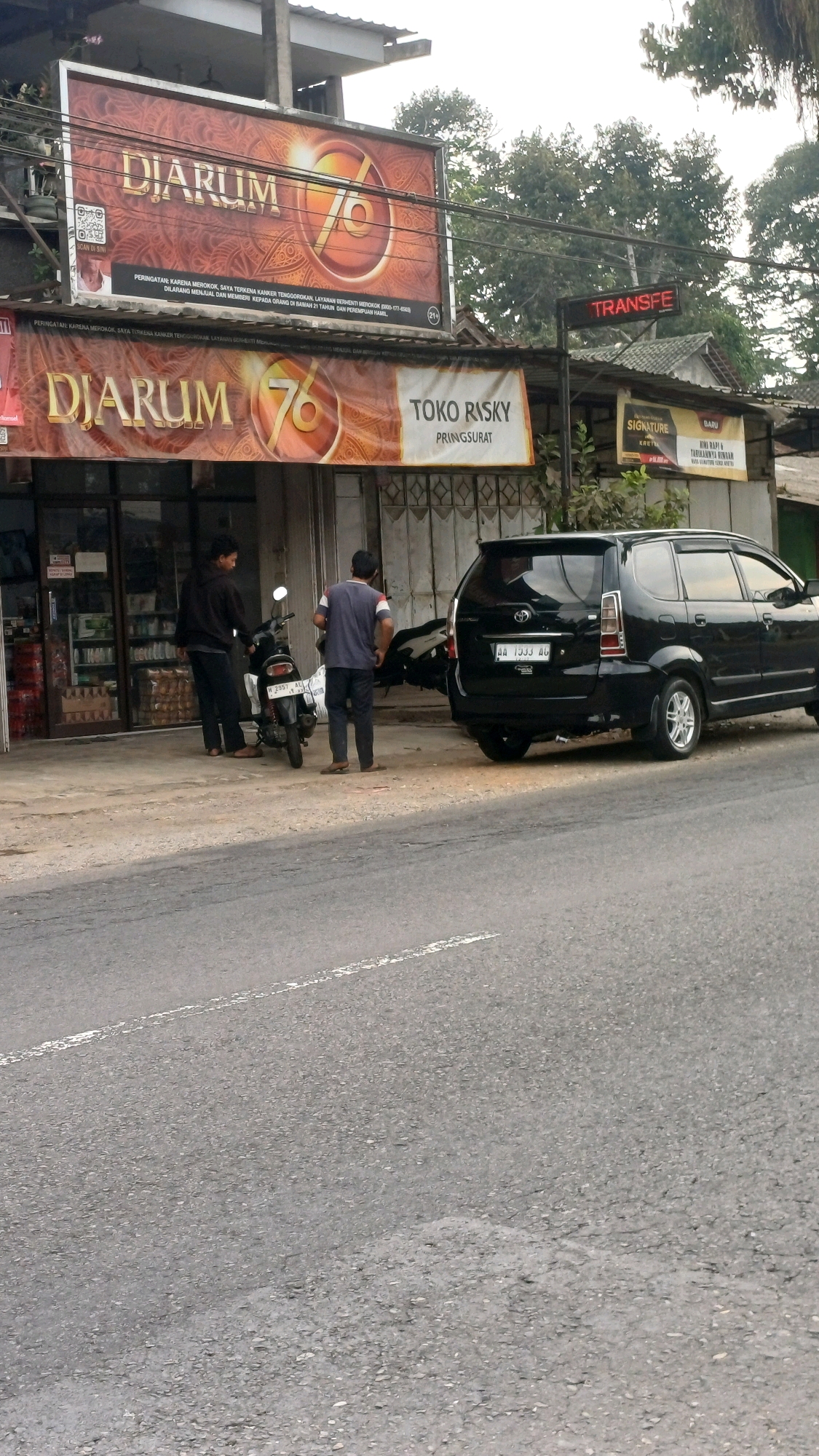 In the city of Tengahan, Indonesia, a street-level view captures a storefront with prominent signage. Above the entrance, a large red and gold billboard displays "DJARUM 76" along with regulatory text in Indonesian concerning smoking and age restrictions. Below this, a smaller banner also features "DJARUM 76" and "TOKO RISKY PRINGSURAT," indicating a shop name and location. To the right of the shop, a red LED sign reads "TRANSFE."
In front of the shop, two individuals stand near a black motorcycle. One person, wearing a dark hoodie and black pants, faces the motorcycle. The other, in a blue t-shirt with white and red accents and dark pants, stands beside the motorcycle, looking towards the right. A black minivan is parked to the right of the motorcycle, facing away from the viewer. Its license plate reads "AA 1933 AG." The ground in front of the storefront is paved, and a grey asphalt road with a white line runs in the foreground. Trees and foliage are visible behind the buildings, suggesting a semi-urban or suburban environment.