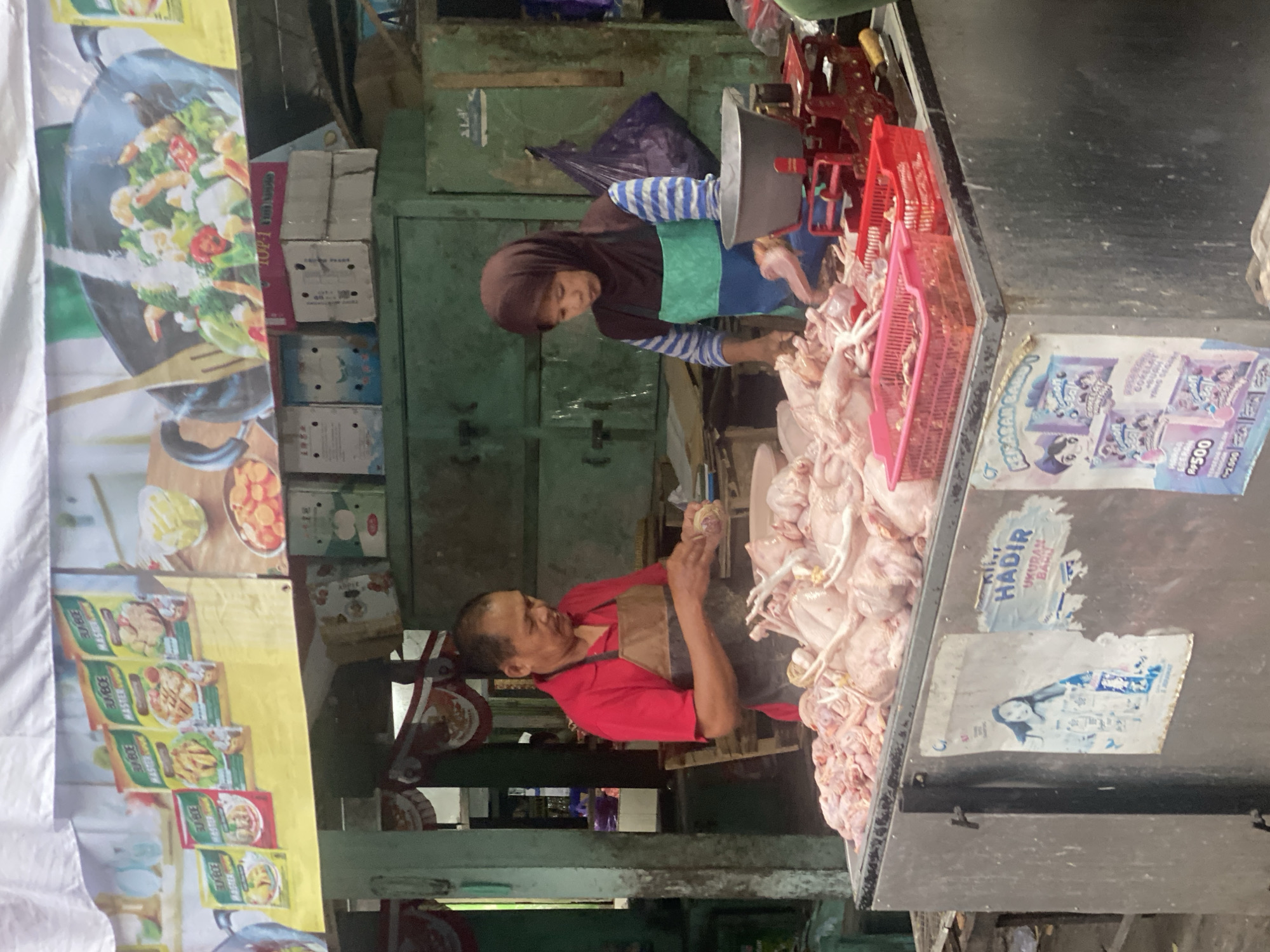 In Ambarketawang, Indonesia, a market scene unfolds as a man and a woman, likely vendors, stand behind a large stainless steel display filled with raw chickens. The man, wearing a red shirt and a brown apron, is actively preparing a chicken. The woman, in a brown headscarf and a patterned apron, is also engaged in processing poultry. A red mechanical scale sits on the counter, accompanied by red plastic baskets containing chicken parts. Behind the vendors, green metal cabinets are visible, suggesting a market stall environment. Banners depicting food products and cooking scenes hang above the stall, adding to the commercial atmosphere. Posters are affixed to the side of the display, featuring images of cartoon characters and pricing information.