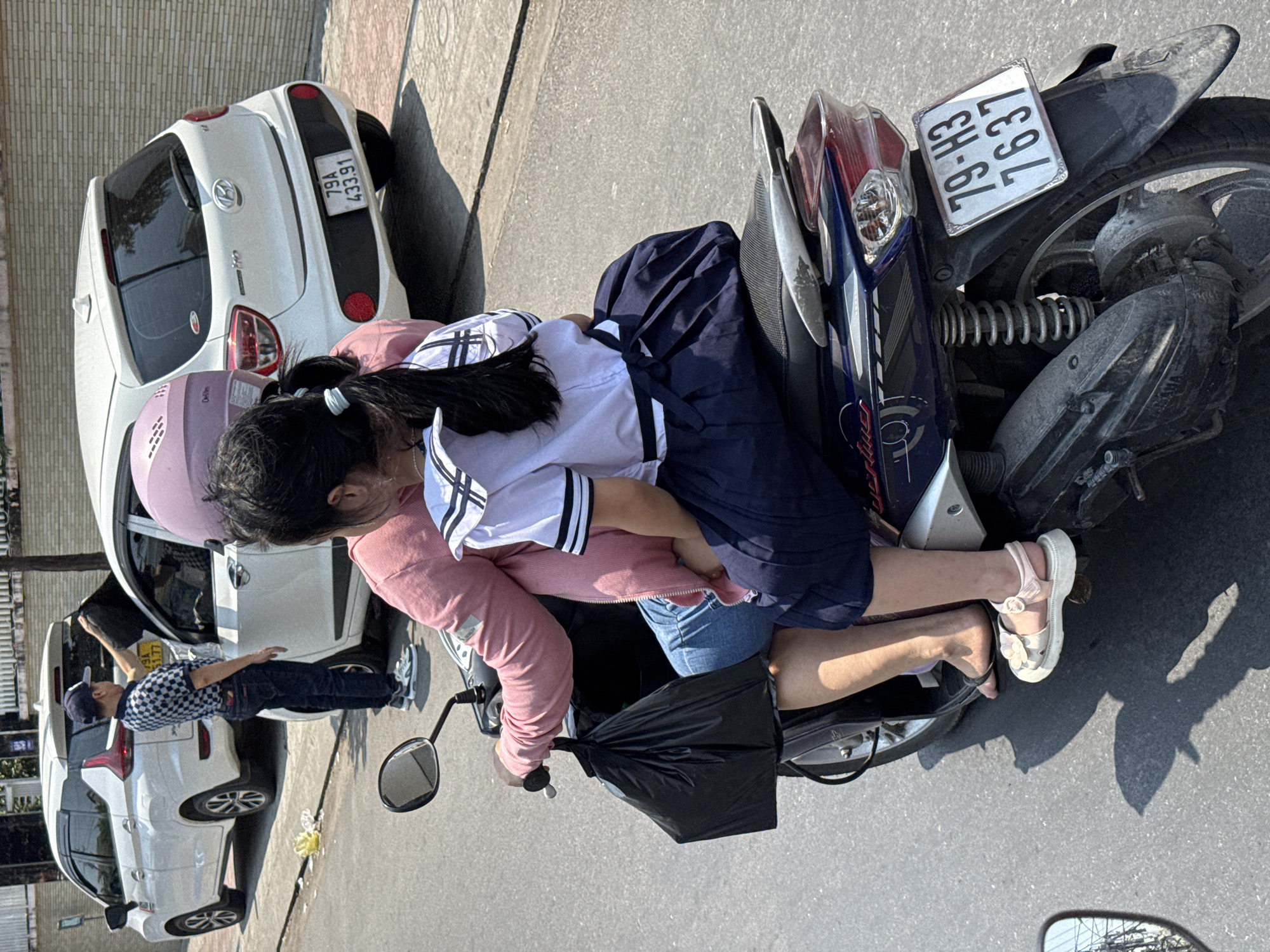 The image captures a scene on a city street. A person wearing a pink helmet and jacket is seated on a motorbike, embracing a younger individual wearing a school uniform with a navy skirt and white top. The motorbike's license plate reads "79-H3 7637." The motorbike is parked adjacent to a white car with the license plate "79A 43391." In the background, another white car is visible, with a person standing next to it wearing a checkered shirt and cap. The environment appears to be a sunny day in an urban setting. The location is in the city of Unknown, Unknown.
