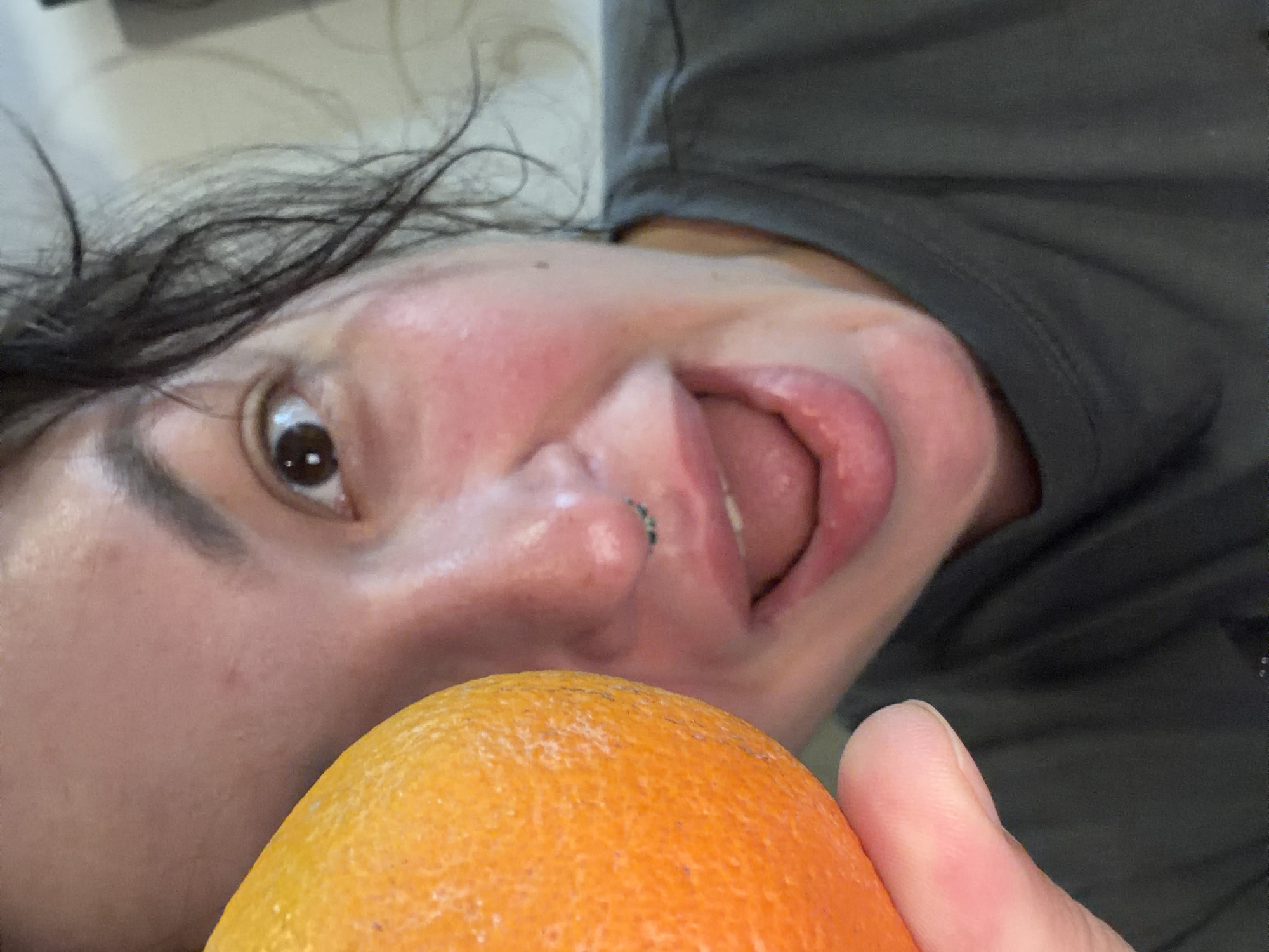 In a close-up, indoor shot, a person's face and an orange are the primary subjects. The person's left cheek, eye, nose, and mouth are visible, along with part of their forehead and hair. They have dark hair, brown eyes, and are wearing a dark gray t-shirt. There is a nose ring on their septum. The person's mouth is open, with their tongue slightly visible. The orange is held close to the left side of the person's face, obscuring a portion of their cheek and forehead. The background is blurred and appears to be indoors, with a light-colored wall visible. There is no discernible connection to Chaikovsky, Russia.