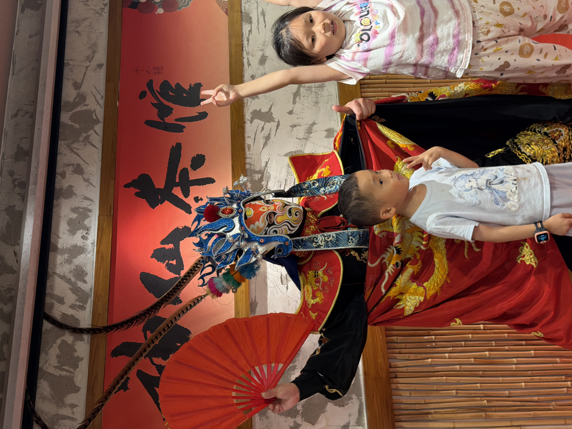 In a scene set in Yuzhong District, China, a person in traditional Chinese opera costume, complete with a decorative headdress and mask, stands holding a red fan. To the right, a young girl poses with her arms raised, making a peace sign with her fingers. To the left and slightly in front of the opera performer, a young boy stands, also making a peace sign. The background features a red banner with Chinese characters and a textured wall. A bamboo-like texture is visible on the lower left side of the image.