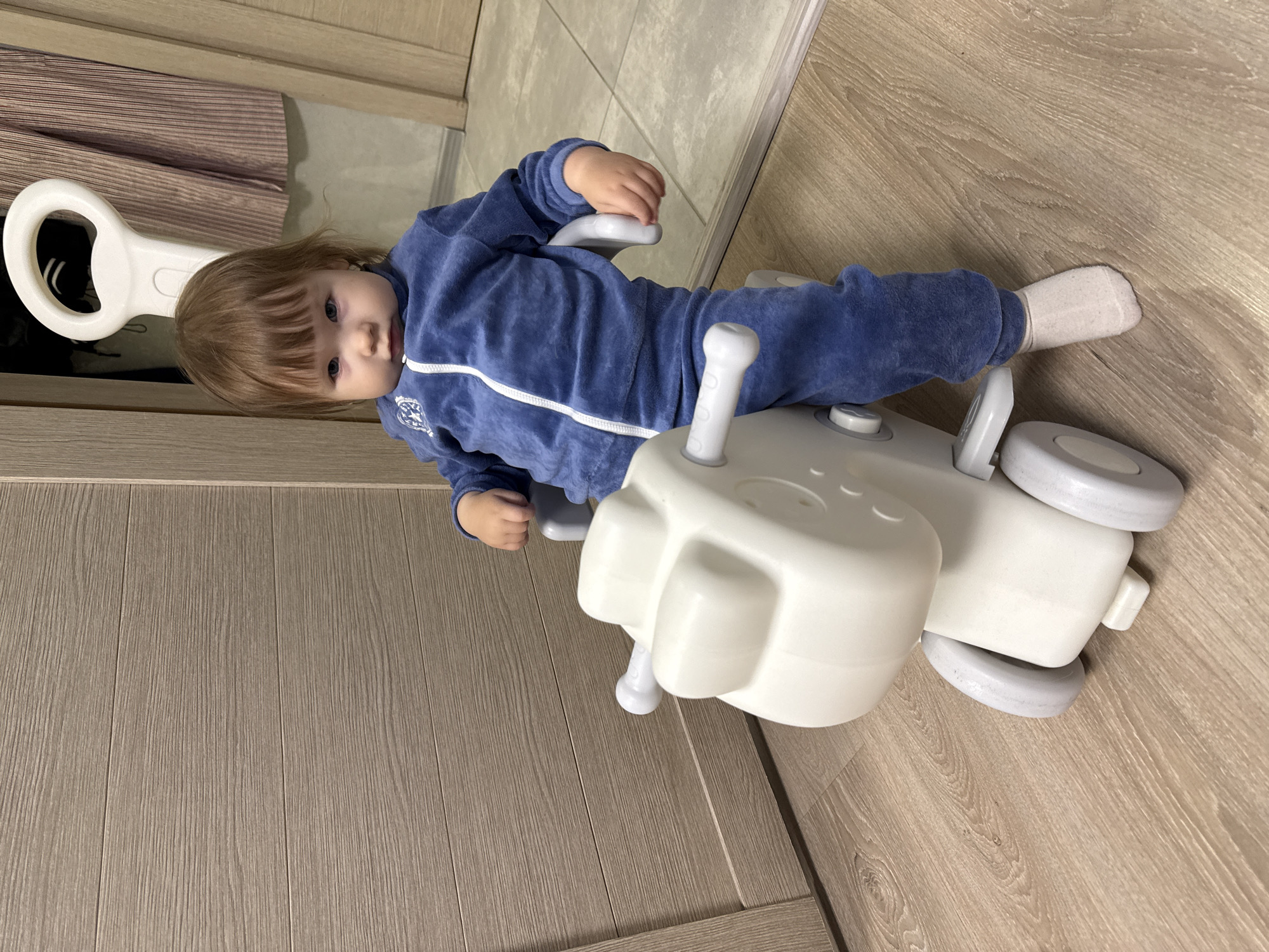 A toddler is sitting on a white toy horse. The toddler is wearing a blue tracksuit and white socks. The toy horse has wheels and a handle at the back. The background shows a wooden door and a ribbed curtain. The floor is made of light-colored laminate. The image appears to have been taken indoors, likely in a residential setting. There is no indication that the scene is in Moscow, Russia.