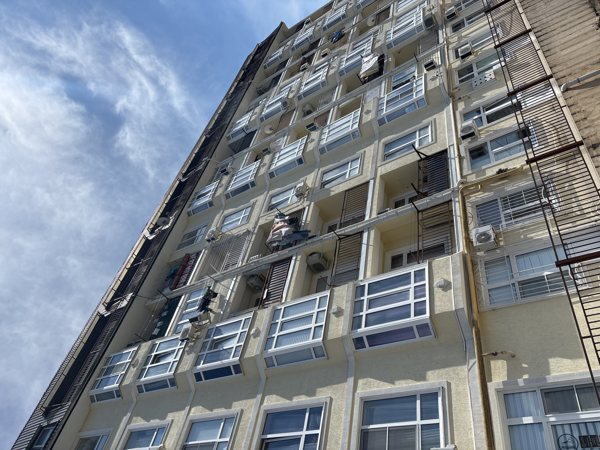 View looking up at the side of a tall apartment building with cream-colored exterior walls. The building has numerous windows, some with white frames, and some are surrounded by air conditioning units. Clothes are hanging from some balconies. A metal fire escape ladder is visible on the right side of the building. The sky is visible above the building with blue patches and scattered white clouds. Based on visual cues, including architectural style and the presence of certain outdoor items, this is plausibly a residential building in the city of Sochi, Russia.