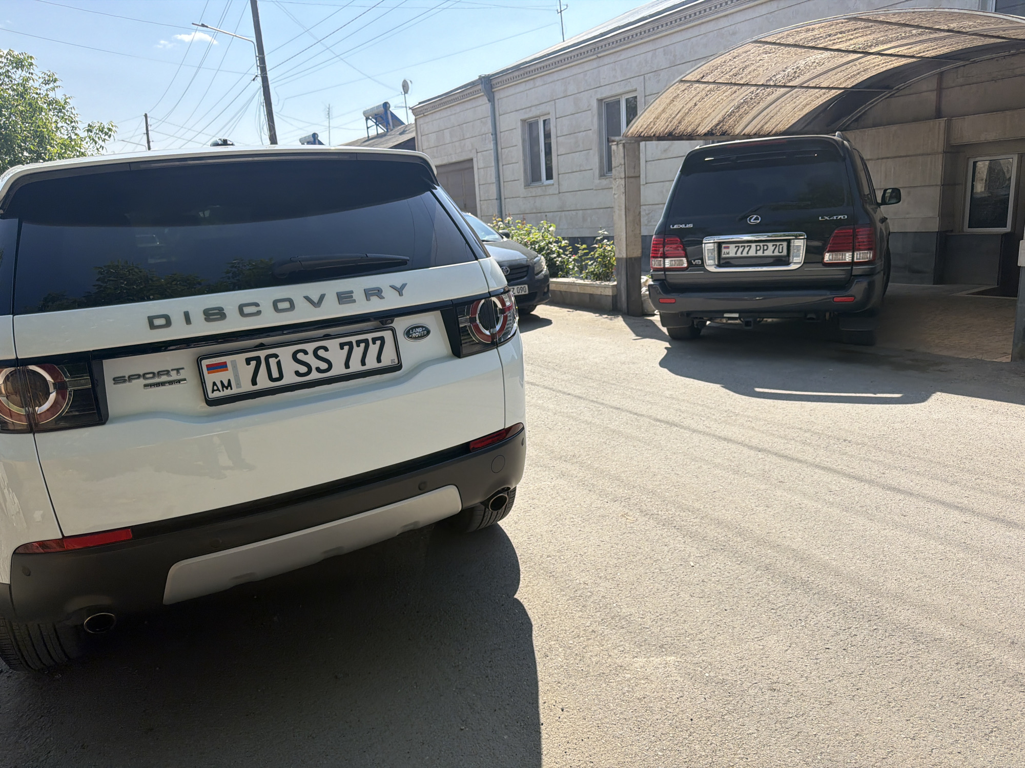 The image shows a street scene with two parked luxury vehicles. In the foreground, there is a white Land Rover Discovery Sport with Armenian license plate "70 SS 777". Behind it, parked under a covered area, is a dark-colored Lexus LX470 also with an Armenian license plate "777 PP 70". A portion of a third car, also dark-colored and with a partial license plate "2 090", is visible between the two SUVs. The scene takes place on an asphalt road in front of a light-colored, one-story building with windows and a covered parking area. Based on the location, it is likely the image was taken in Dimitrov, Armenia.