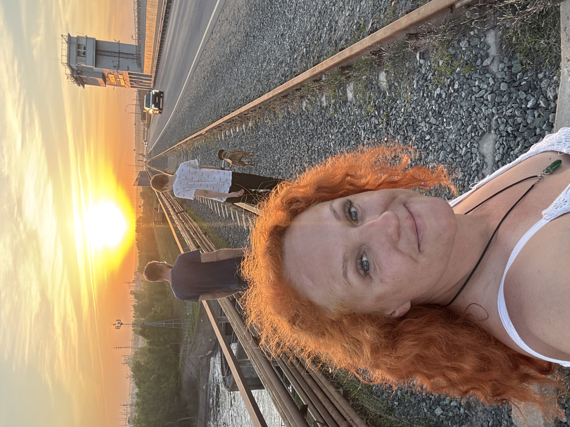 The image captures a selfie of a woman with red hair, framed against a sunset. She is wearing a white tank top and a necklace with a green pendant. Behind her, two men are walking along a set of railroad tracks, one of them with a dog. In the distance, a tall industrial structure is visible to the left of the setting sun. To the right of the industrial structure is a road with a car on it.
The location is identifiable as Chaikovsky, Russia, based on the backdrop of industrial infrastructure typical of the area and the distinctive architectural style of the tall building in the background. The scene depicts a recreational activity with a sunset backdrop, contrasted with industrial elements in the environment.