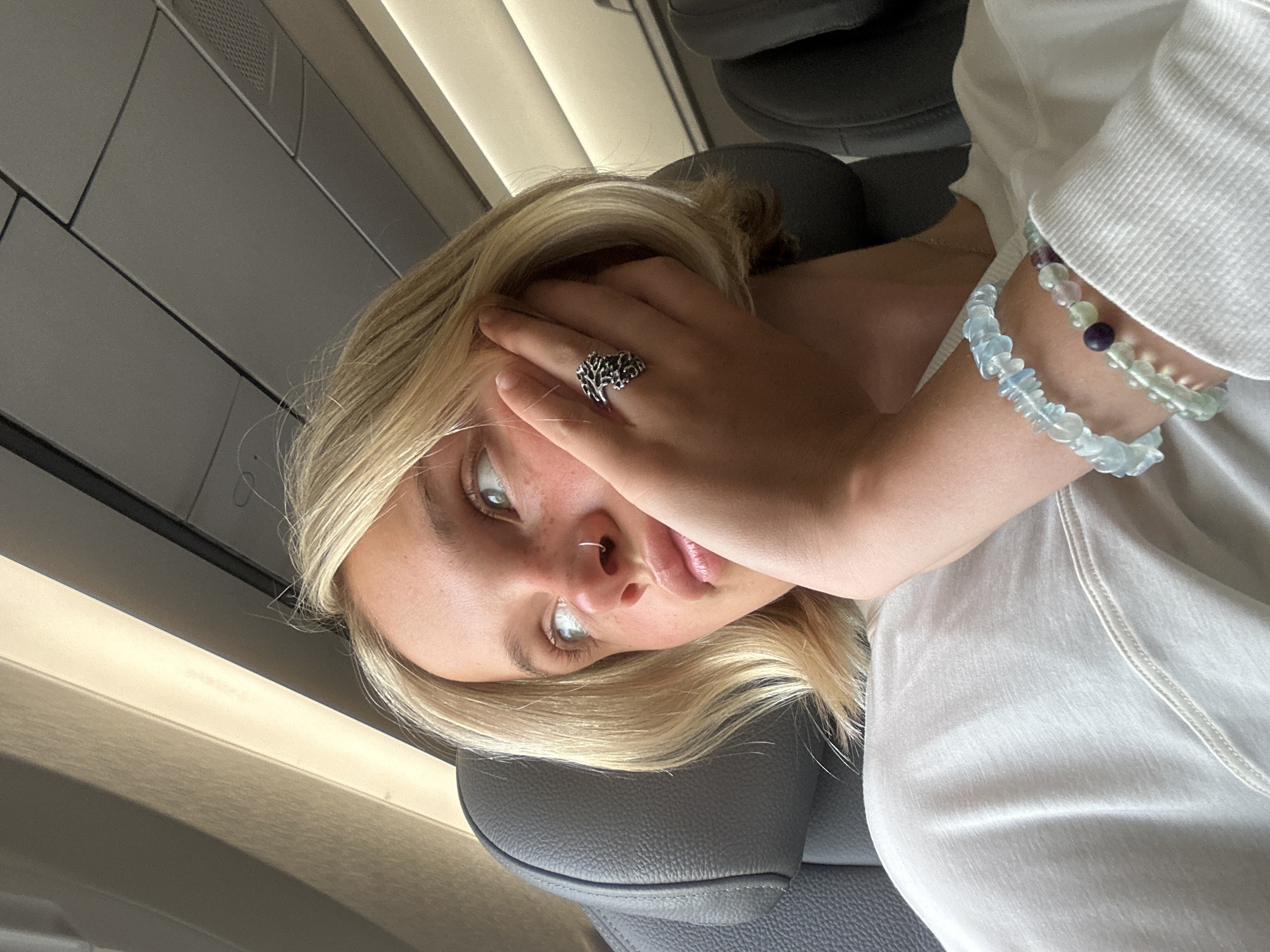 A young person with short blonde hair and freckles is seated on an airplane, resting their chin on their hand. They are wearing a white t-shirt, a ring on their right hand, and two beaded bracelets on their left wrist. The lighting suggests daytime, with natural light filtering in from a window. The background shows airplane seating and overhead compartments. The location is inside an airplane cabin, which could be at Dorval, Canada, if the flight originated or is destined for Montréal–Trudeau International Airport (YUL).