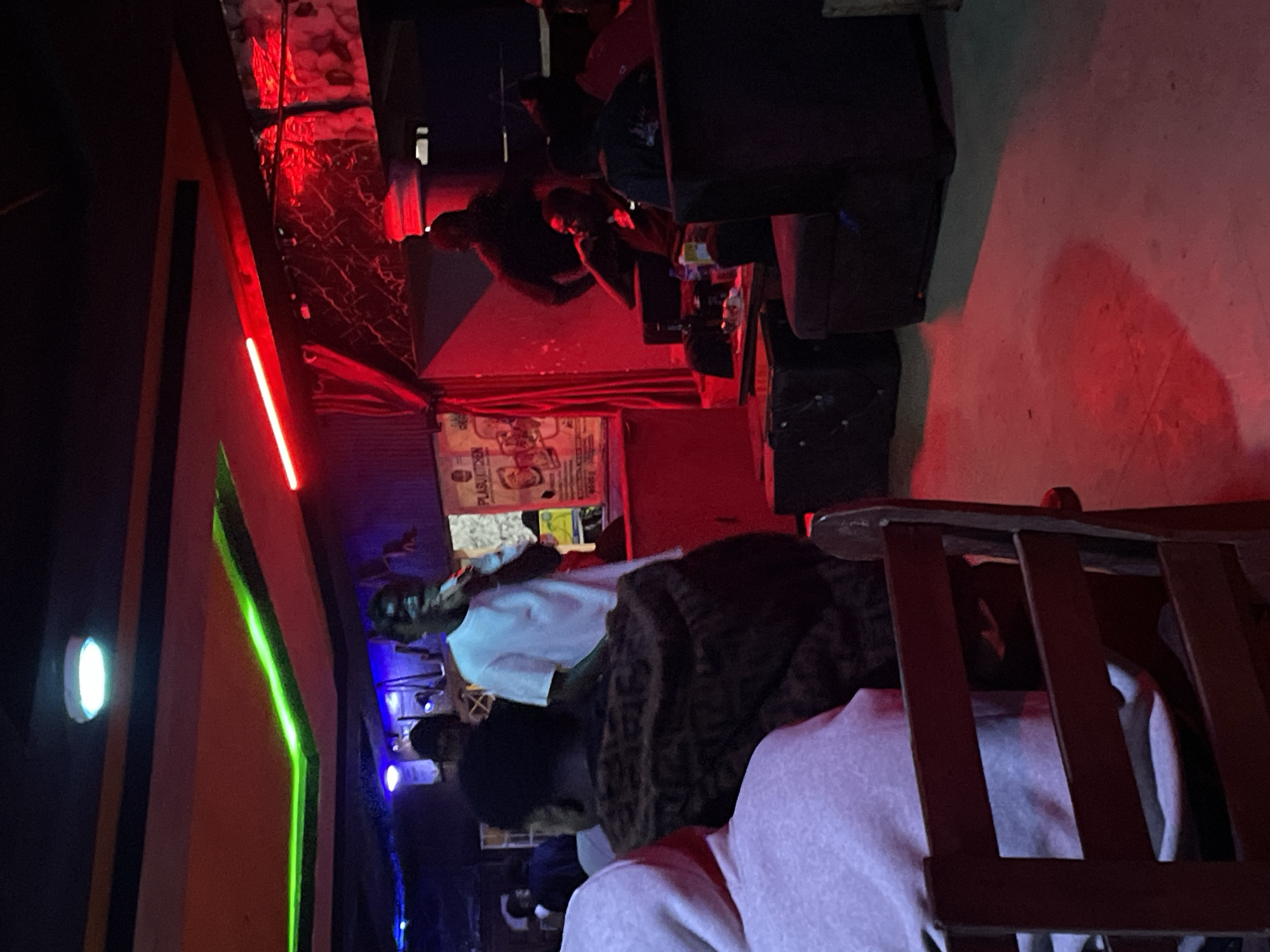 In Durumi, Nigeria, an indoor venue, possibly a bar or club, is dimly lit with red and green neon lights. A man in a white t-shirt stands at a podium, holding a microphone, appearing to perform or speak to the audience. Several individuals are seated in plush seating areas, some with drinks on tables. The atmosphere suggests a lively evening event.