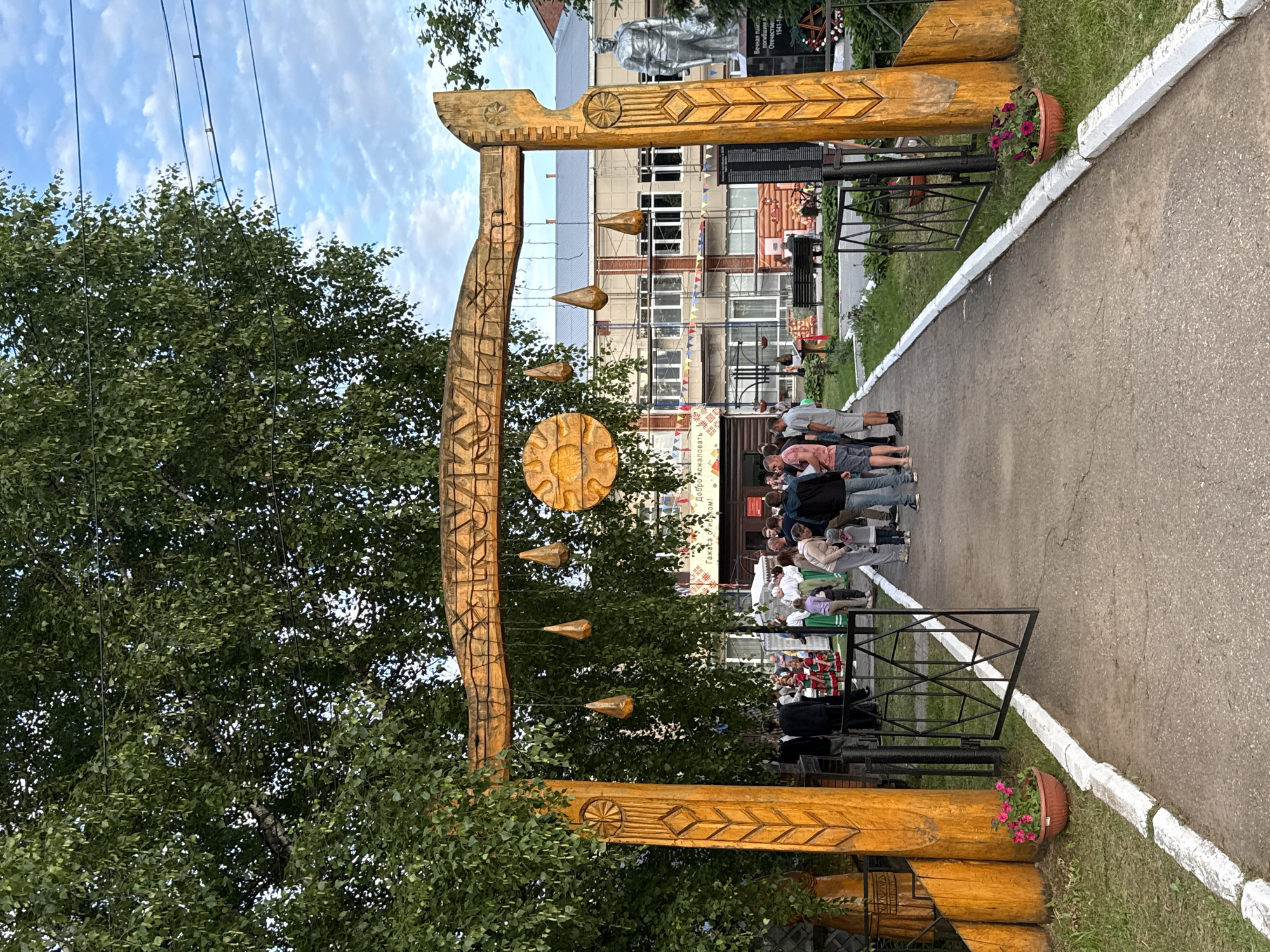 An outdoor scene in the city of Kukui, Russia, features a large, ornately carved wooden archway with traditional Russian folk art motifs. The archway is adorned with decorative wooden elements and hanging tear-drop shaped ornaments. A central circular sun-like carving is also visible on the arch. A crowd of people, dressed in casual attire and some in traditional clothing, are gathered in the pathway leading through the arch. In the background, a multi-story building with a light-colored facade and numerous windows is visible. To the right of the archway, there is a memorial with a statue of a soldier and an inscription. The ground is paved with asphalt, bordered by a white-painted curb and grassy areas with flower pots. Trees with dense green foliage are present, and the sky is partly cloudy.