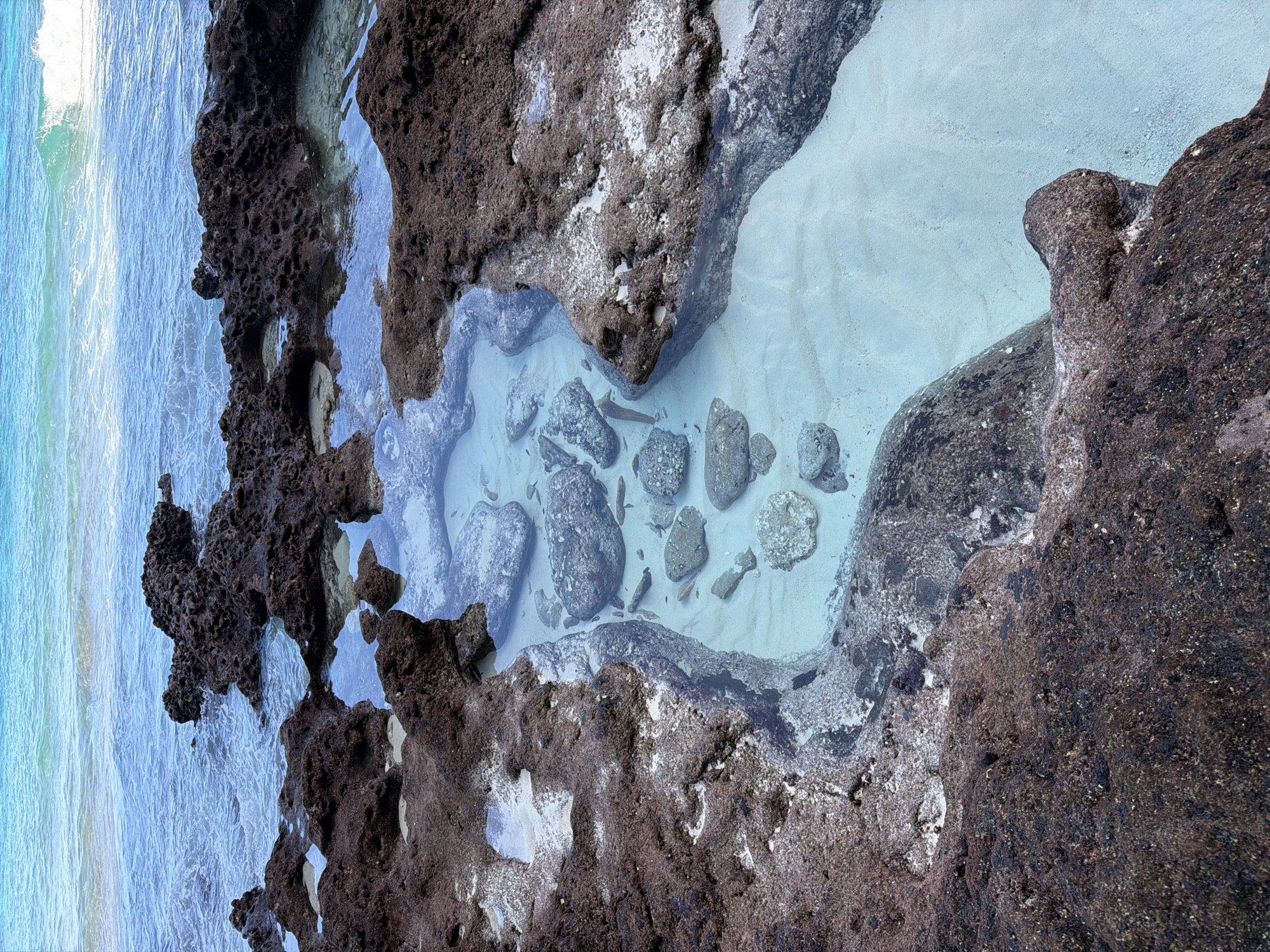 The image shows a rocky tide pool in Pejukutan, Indonesia. The pool is formed by dark, porous rocks covered in what appears to be moss or algae. The water is clear and shallow, revealing a sandy bottom with several smooth, grey rocks of various sizes scattered within. The ocean's edge is visible on the left, with blue-green water and small white waves.