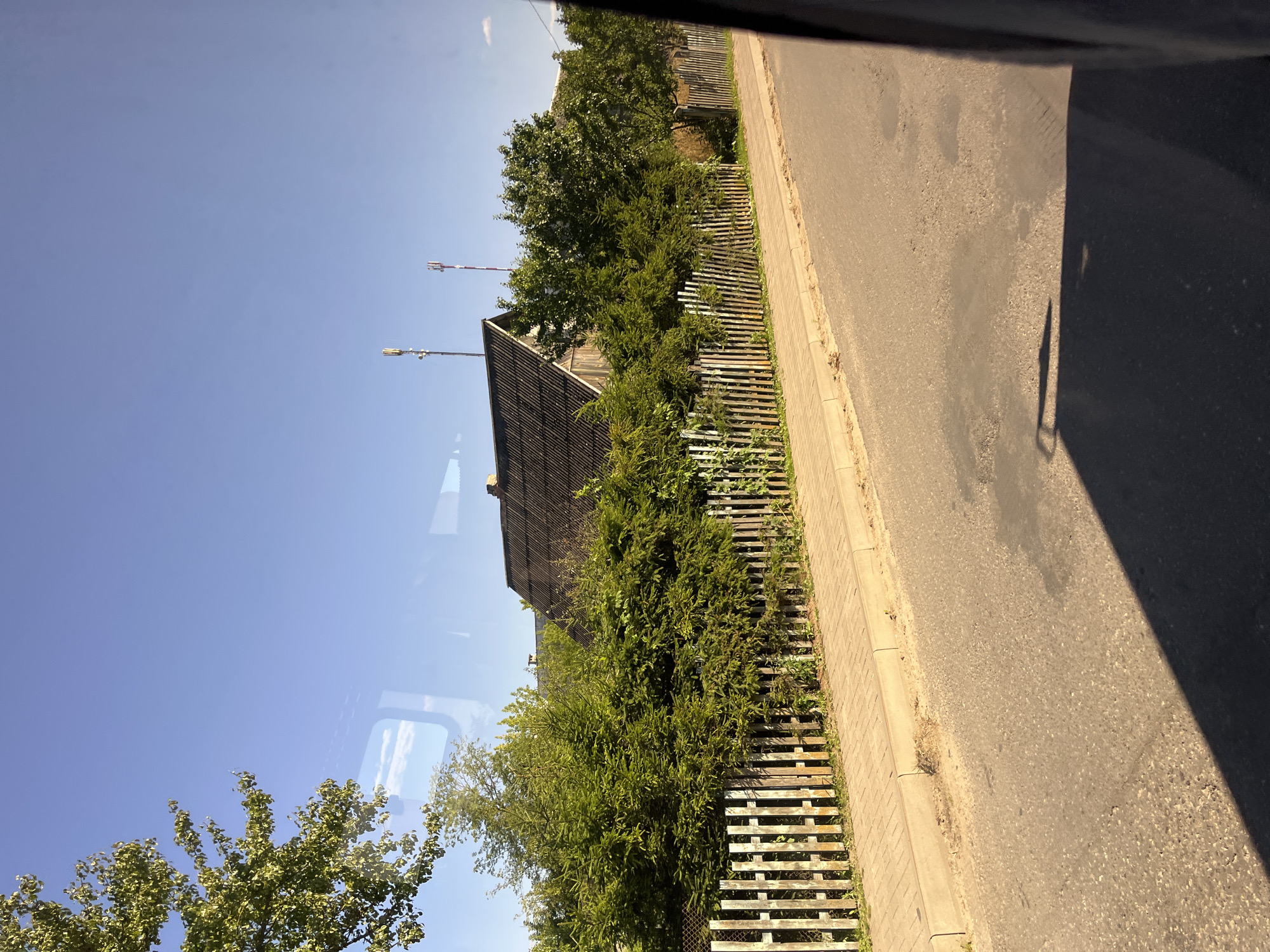 The image captures a view from a vehicle, likely a bus or car, looking out at a rural or suburban street scene in Pastavy, Belarus. The foreground features an asphalt road with some patches and a shadow cast across it, suggesting it is daytime and sunny. To the left of the road is a paved sidewalk or pathway with a light-colored curb. Beyond the sidewalk, a wooden fence with vertical planks runs along the property line.
Behind the fence, a dense line of evergreen trees and bushes forms a natural barrier. Peeking through the foliage are parts of a house with a dark, corrugated roof and wooden walls. Two communication towers are visible in the distance above the trees, indicating some degree of modernization in the area. The sky is a clear, bright blue. The overall impression is one of a quiet, sunlit day in a residential area.