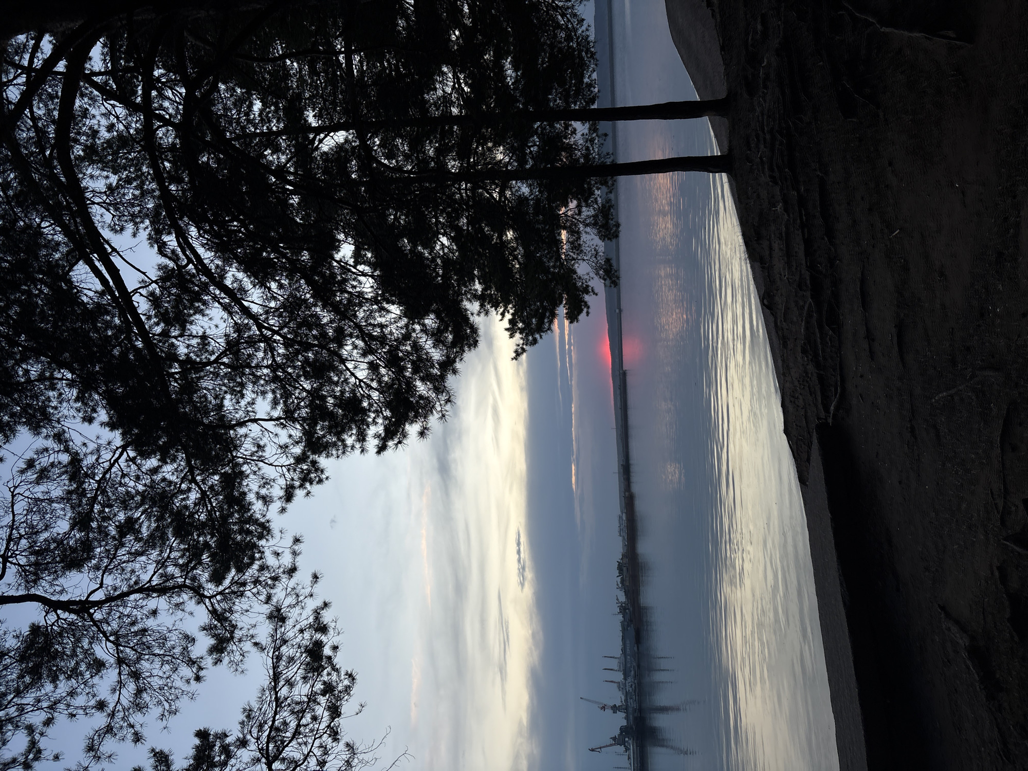 The image shows a body of water, likely a river or reservoir, with a visible skyline in the distance, likely depicting a port or industrial area. Silhouetted trees frame the left side of the frame, adding depth. A small, concentrated red light is visible on the horizon, suggestive of a sunrise or sunset partially obscured by clouds. The shoreline is visible to the right, displaying vegetation and earth. Given the reference to Chaikovsky, Russia, and the landscape elements, it is possible the location is near the Kama River or the Votkinsk Reservoir. However, without more specific details, it is not possible to pinpoint the exact location. The image appears to be taken during twilight or dawn hours. No individuals are apparent.