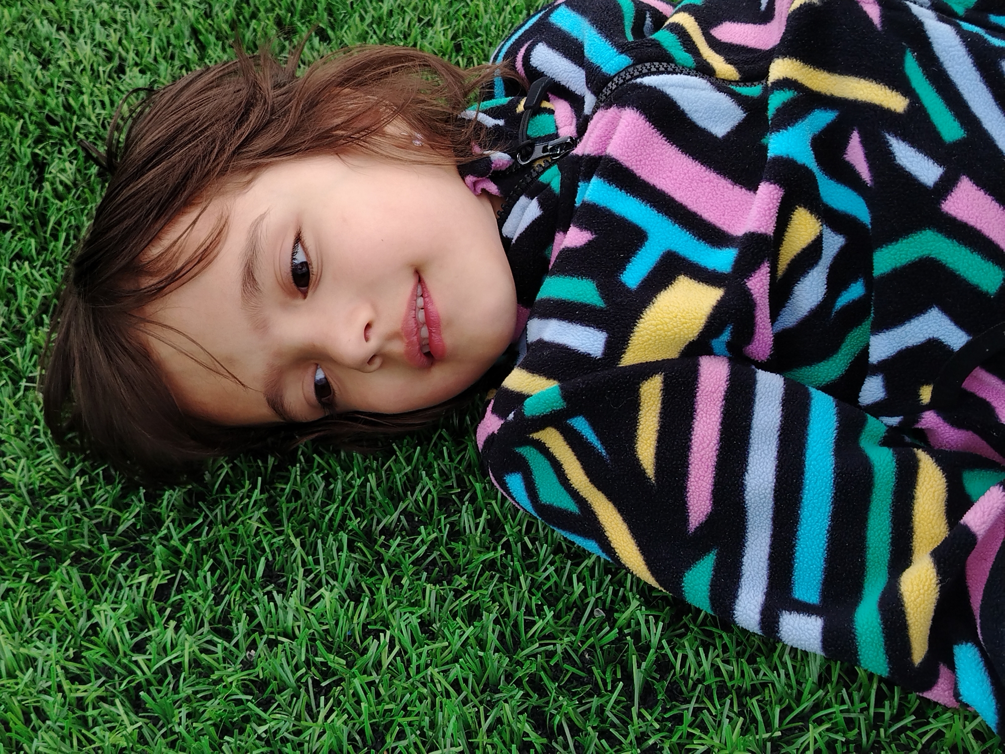 A young child with brown hair is lying on artificial green turf. The child is wearing a black fleece jacket with a colorful geometric pattern, including pink, turquoise, yellow, and green. The child is looking toward the camera with a slight smile. The background consists entirely of the artificial green turf. There is no indication of the scene being in Noviy, Russia.