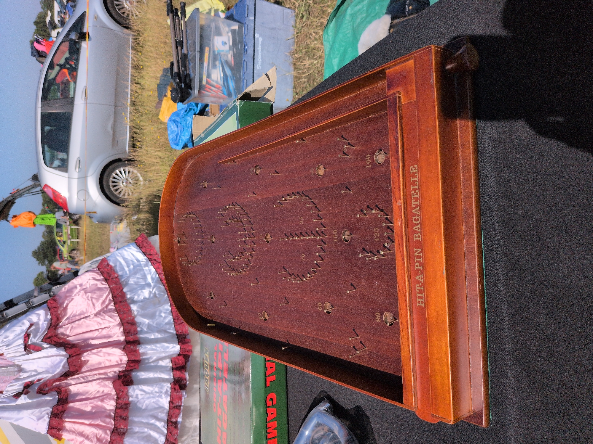 The image depicts a vintage wooden bagatelle game set on a black surface, likely a table or stall at an outdoor market. The game board is made of polished wood and features a series of brass pins and several circular holes with numerical scoring values printed around them, including 50, 60, 70, 90, 100, 125, and 150. The text "HIT-A-PIN BAGATELLE" is inscribed on the front of the wooden frame.
In the background, a large, ruffled pink and white dress is visible, suggesting a costume or theatrical prop. Behind the dress, a silver car is parked on a grassy area. Further in the distance, other vehicles and people can be seen, as well as trees and a bright blue sky, indicating an outdoor setting. A green box with red lettering that appears to say "JACQUES" and "AL GAM" is positioned to the left of the bagatelle game. Other items, including what appears to be a blue tarp, a clear plastic container with various objects, and a tripod, are also present in the background.
The overall scene suggests a fair, fete, or car boot sale in an open, possibly rural or suburban environment. While the exact location within Claverley, United Kingdom is not explicitly identifiable from the visual elements, the context of an outdoor market is strongly suggested.