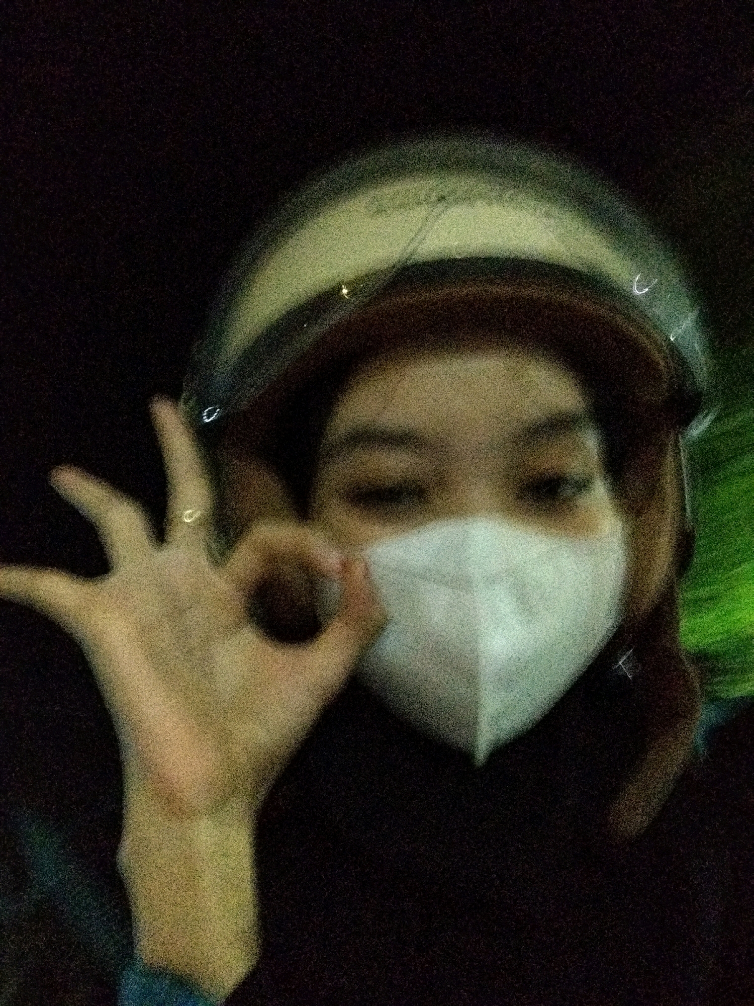 A person, wearing a helmet and a white face mask, is captured in a blurry, dimly lit, close-up shot. Their right hand is held up, forming an "okay" gesture with the thumb and index finger touching, and a ring is visible on the ring finger. The background is dark, with streaks of light suggesting movement, possibly from a vehicle. There is no discernible location in Semanding, Indonesia.