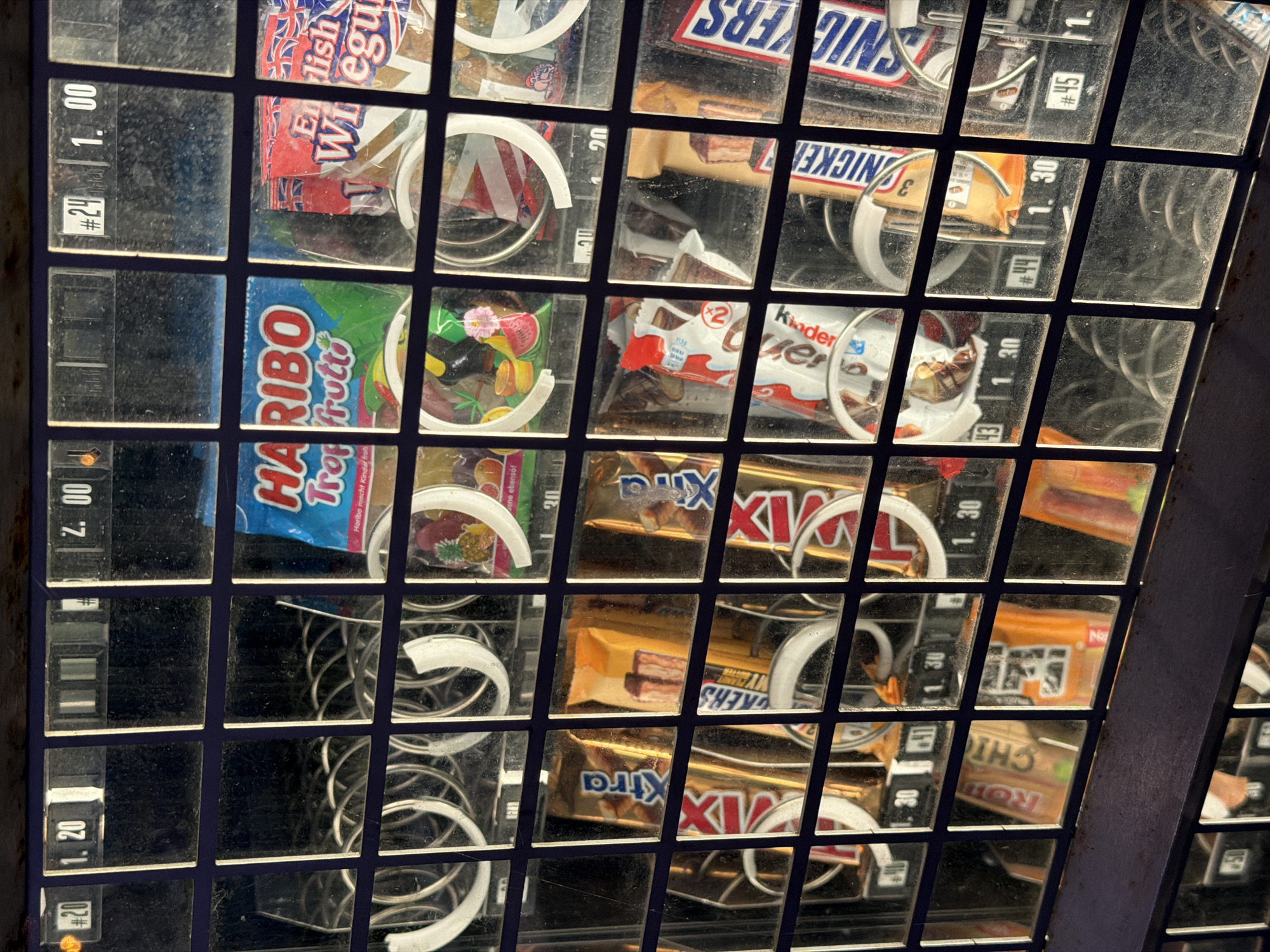 The image shows a section of a vending machine filled with various candy bars and bags of sweets, likely located in Witten, Germany, as indicated by the prices displayed in Euros. The machine has a grid of glass compartments, each containing a product and a corresponding price label.
In the top row, visible products include a bag of Haribo Trop Froot, and two bags of what appear to be English sweets. Prices shown are 1.20, 2.00, and 1.00.
The second row displays several chocolate bars. Brands identifiable include "Xtra," and "Kinder" (likely Kinder Chocolate). Prices for this row range from 1.30.
The third row also contains candy bars, including what appears to be Snickers bars. Prices are consistently 1.30 in this section.
The vending machine has a metallic outer frame and a dark blue background behind the products. The overall condition of the machine appears to be functional, though the glass displays show some dust and smudges. No people are present in the image. The lighting suggests it might be daytime, possibly with artificial light from within the machine.