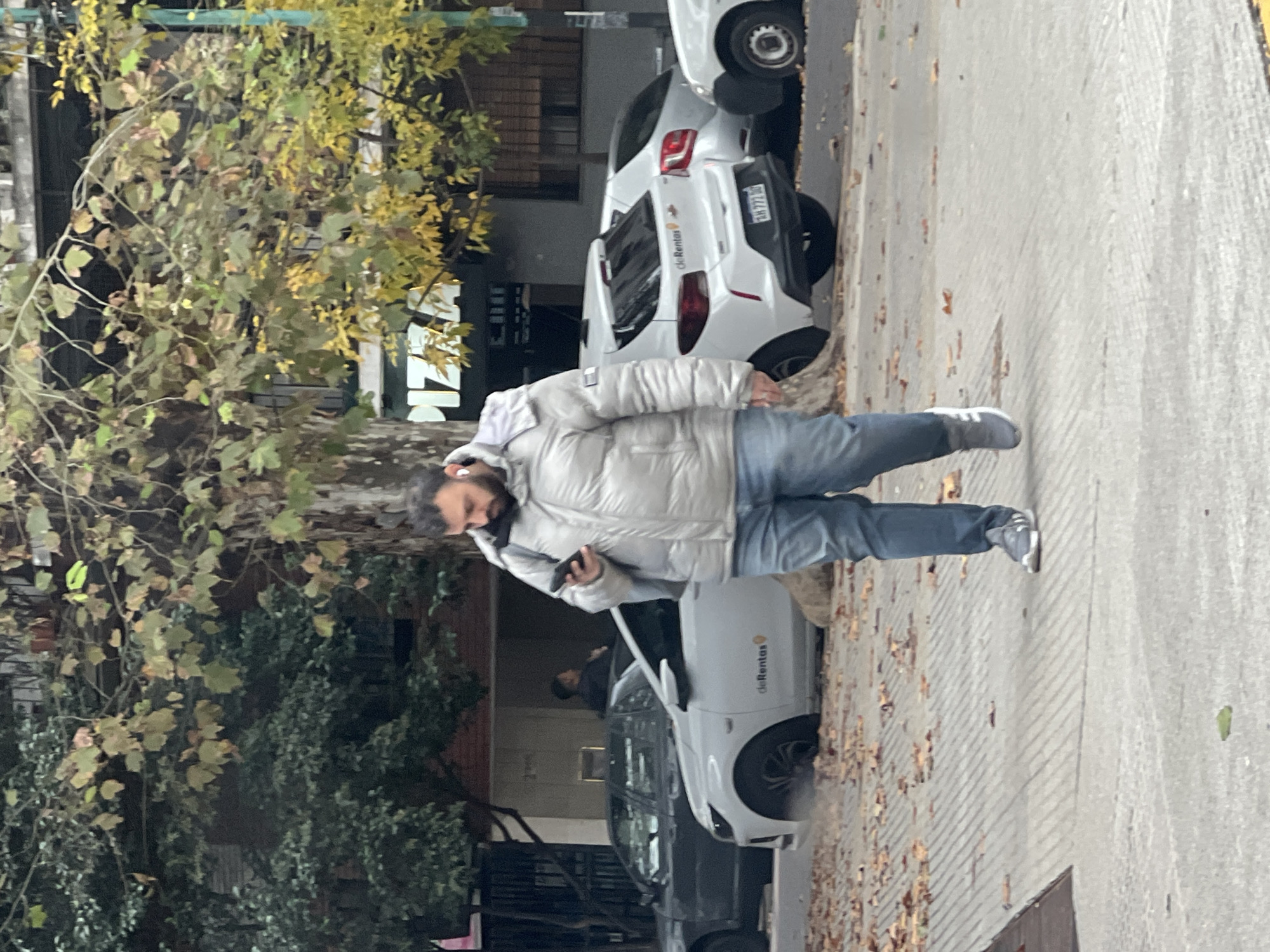 A man with a beard, wearing a white puffy jacket, light blue jeans, and grey sneakers, walks on a sidewalk while looking at his mobile phone. The sidewalk is covered in fallen leaves. Behind the man, there are several cars parked. The cars are of the brand De Rentas. A tree with yellowing leaves is visible to the left of the man. The location is the city of Buenos Aires, Argentina.