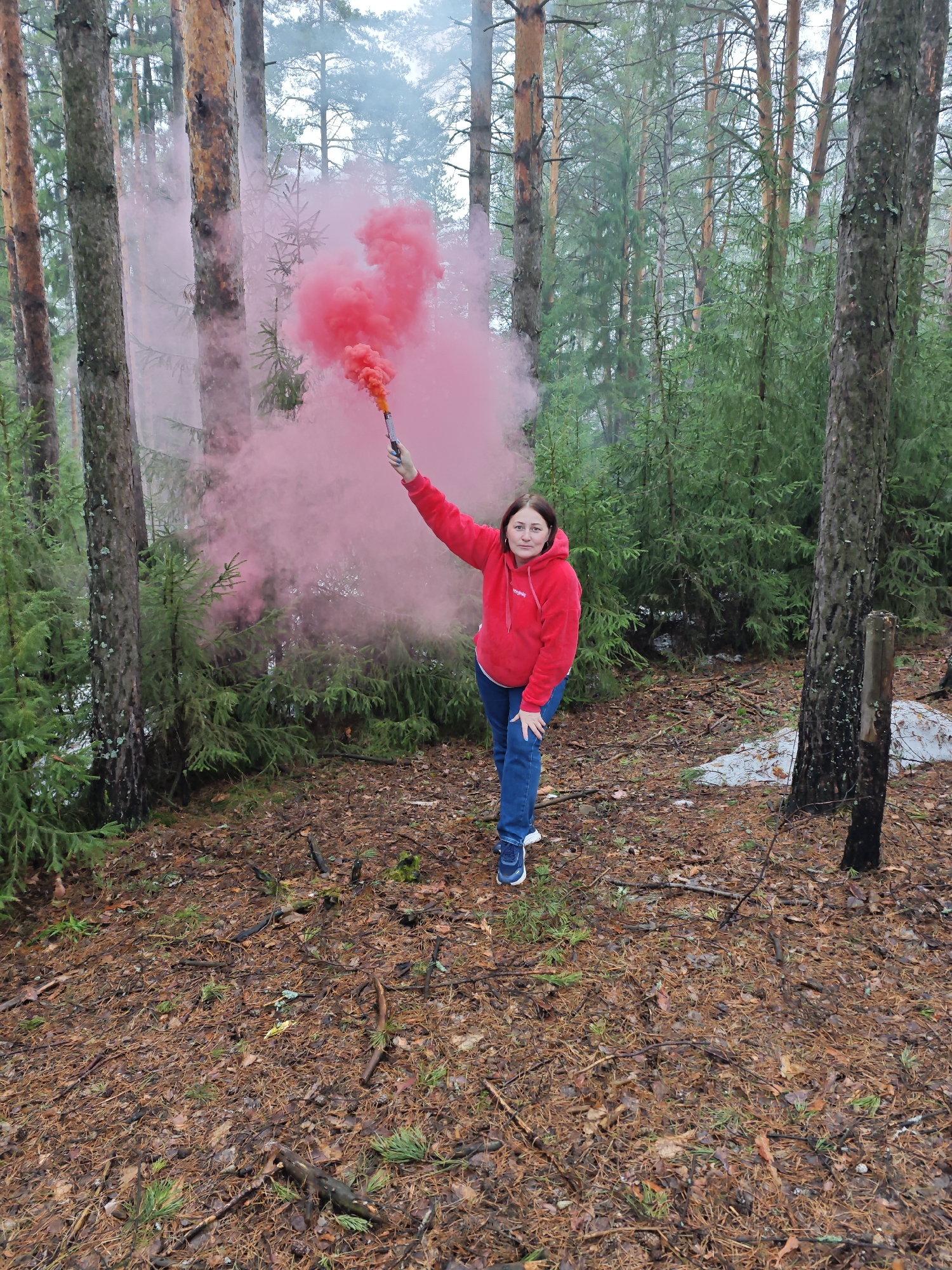 A woman stands in a forest, holding a smoking red flare in her right hand. She is facing the camera, wearing a red hooded sweatshirt and blue jeans, with sneakers visible on her feet. The location is a forest with tall pine trees and smaller evergreen trees. The ground is covered in pine needles and some vegetation. There is partial snow cover in the background. The scene appears to be set in Марковский, Russia, based on provided location context.