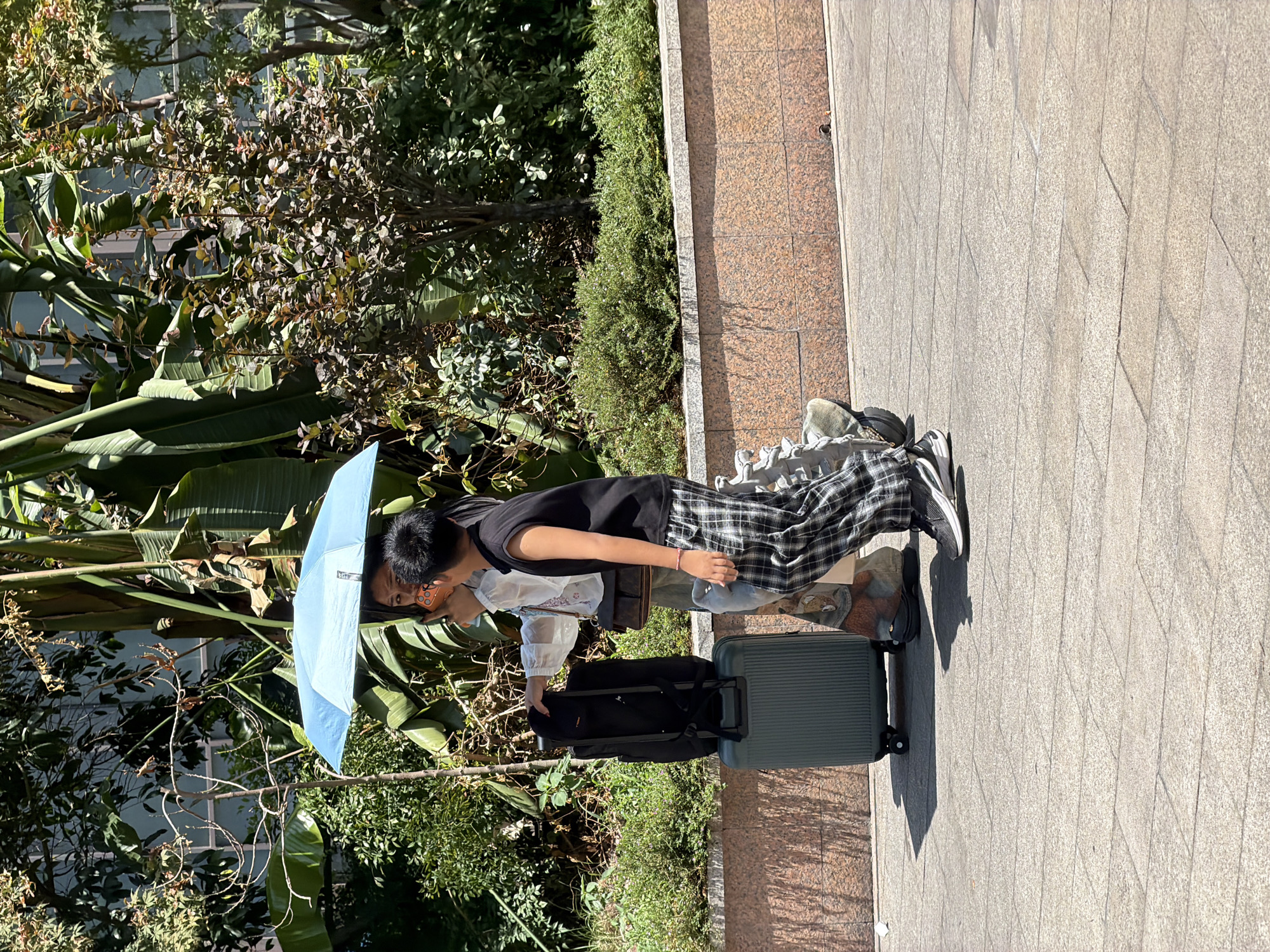 Two individuals, a woman and a boy, walk on a paved walkway in what appears to be a park or garden area. The woman, on the left, is holding a light blue umbrella over both of them, shielding them from the sun. She is wearing a white, sheer blouse and patterned pants, and is looking at her phone, which she is holding up. A large, dark suitcase and a black backpack are positioned next to her. The boy, on the right, is wearing a black sleeveless shirt and plaid pants, and is looking ahead. They are walking towards the right side of the frame. The background consists of lush green foliage, including large-leafed plants and smaller bushes. A stone wall runs horizontally behind the walkway, with a textured, reddish-brown surface. The ground is made of light-colored rectangular pavers. The lighting suggests it is daytime, with harsh shadows cast on the ground. The overall setting suggests a sunny day in an urban green space, possibly in the Yuzhong District of China, given the context.