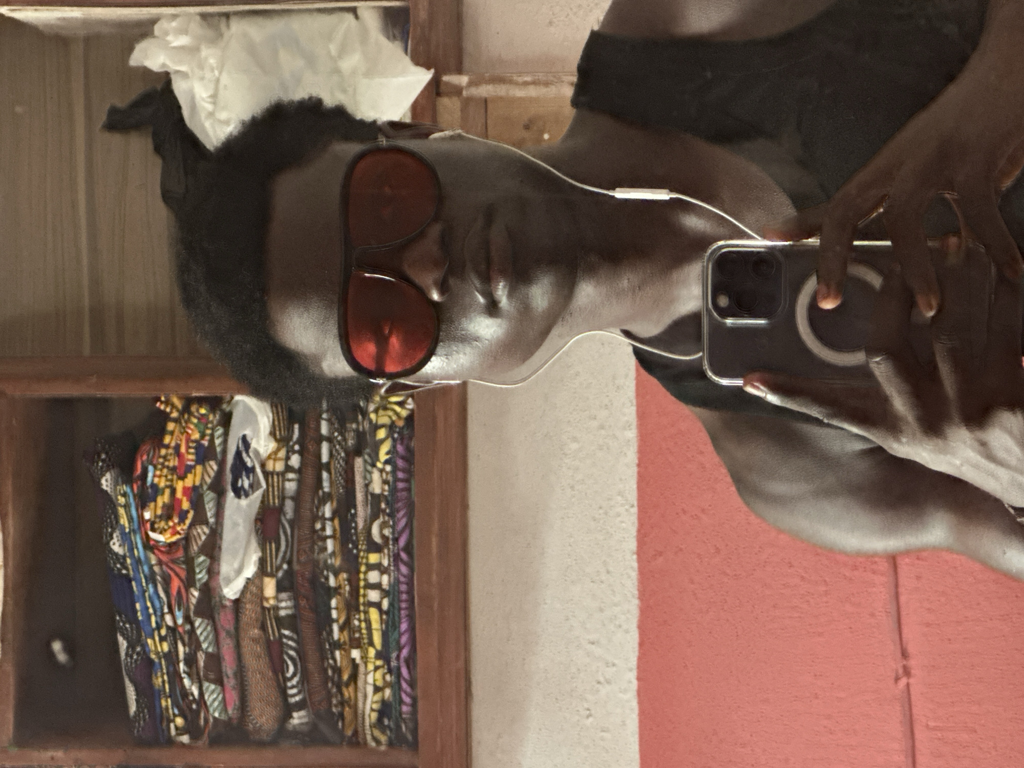 An individual with dark skin, wearing a dark top, red-tinted aviator-style sunglasses, and wired white earphones, is captured in a mirror selfie. Their reflection shows them holding a smartphone with a clear case that features a MagSafe-like ring, indicating they are taking the photo themselves. The person's expression is neutral and calm.
The setting appears to be indoors, possibly a room or shop in Badagry, Nigeria, given the distinctive patterned fabrics. To the left of the individual is a wooden shelving unit or cupboard. The top shelf contains a white crumpled item, possibly tissue paper or a piece of cloth. Below, several vibrant, patterned fabrics, likely traditional African textiles such as Ankara prints, are neatly folded and stacked.
The background also features a wall with two distinct sections: one with a light, rough texture and another with a reddish-orange, rough texture. The overall lighting suggests an indoor environment, possibly with artificial light, casting some soft shadows. There is no visible text in the image.